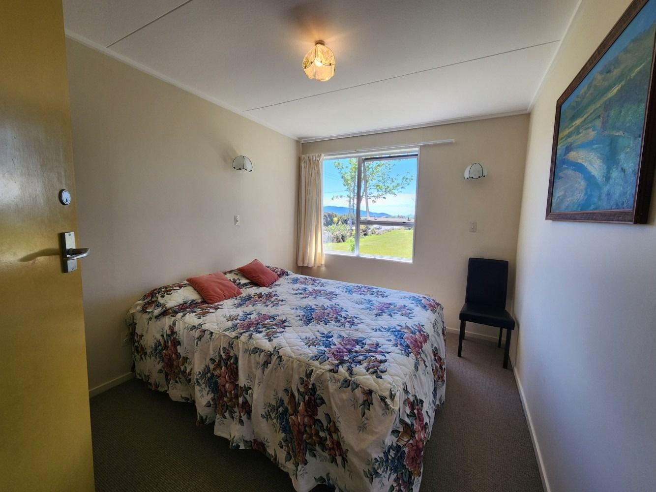 Bedroom with a double bed, floral bedspread, window, artwork, and chair.