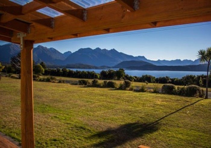 Wooden pergola frames a scenic view of a lake, grassy field, and mountain range under a clear blue sky.