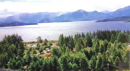 Lakeside view with evergreen trees in the foreground, campsite, and mountains in the distance under a cloudy sky.