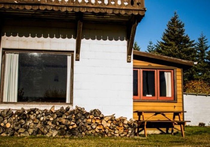 A white and yellow cabin with stacked firewood and a picnic table outside on a sunny day.