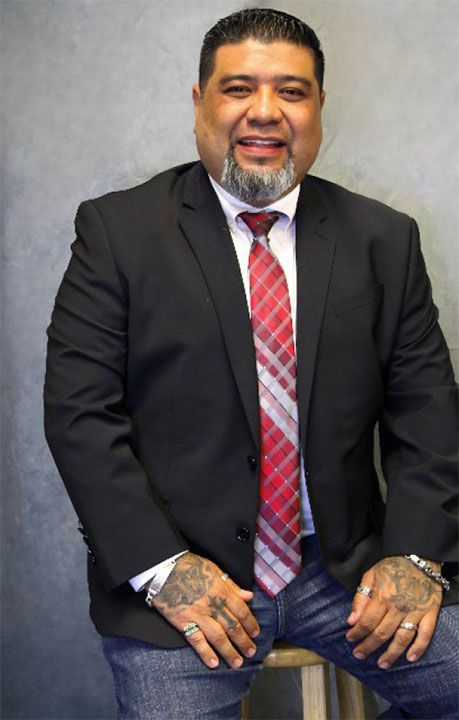 Man with tattoos wearing a suit and tie, smiling, leaning on a stool. Gray background.