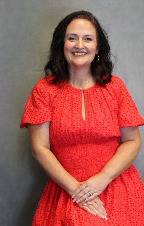 Woman in a red eyelet dress smiles, hands clasped, against a gray wall.