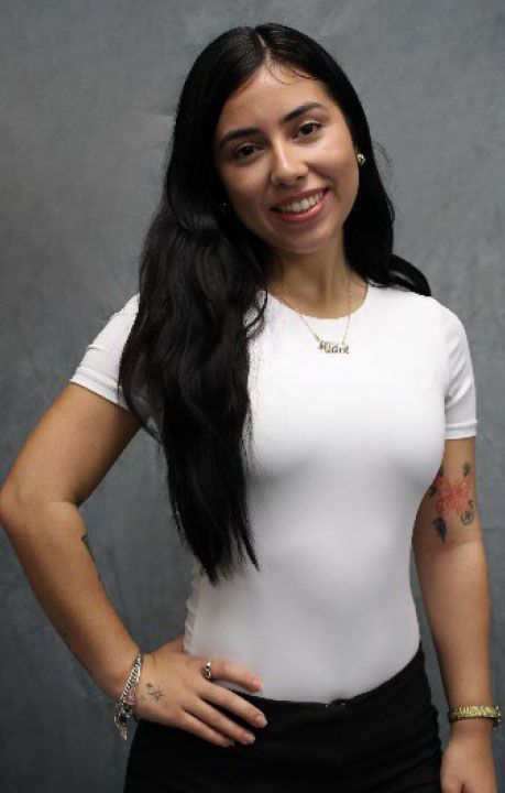 Woman with dark hair smiles, wearing white top and black pants, arm on hip, tattoo on arm.