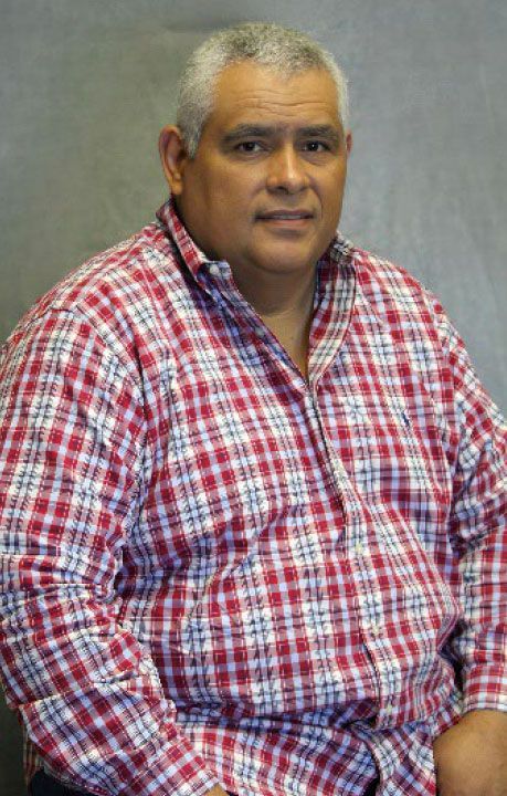 Man in a red and white plaid shirt, seated, looking at the camera. Gray hair, neutral background.
