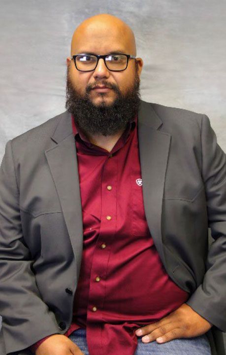 Man with glasses, shaved head, and beard wearing a red shirt and gray blazer, seated.