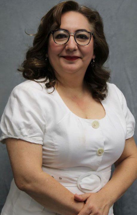 Woman with glasses, smiling, wearing a white dress. Gray background.