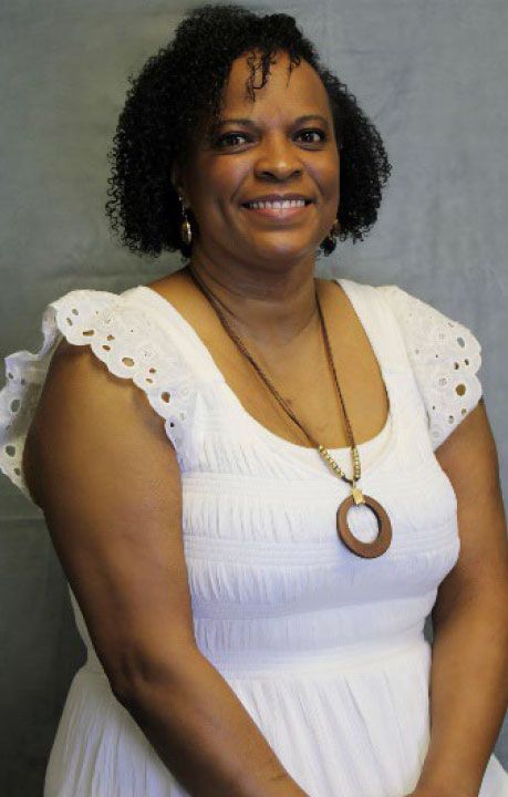Woman with curly hair, wearing a white dress, smiling, and a wooden pendant.