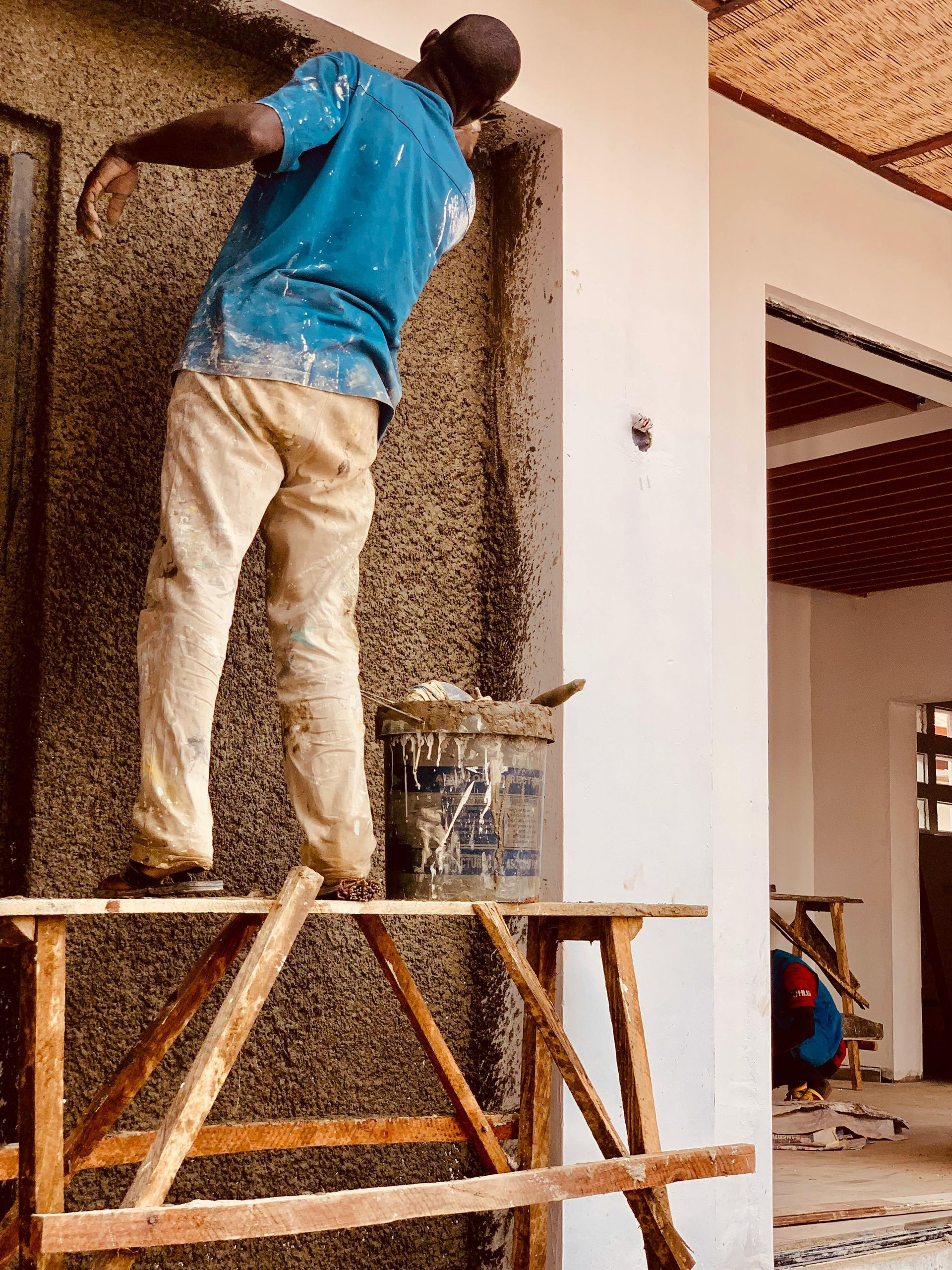 Man plastering a wall while standing on a wooden scaffold outdoors. He's wearing a blue shirt and beige pants.