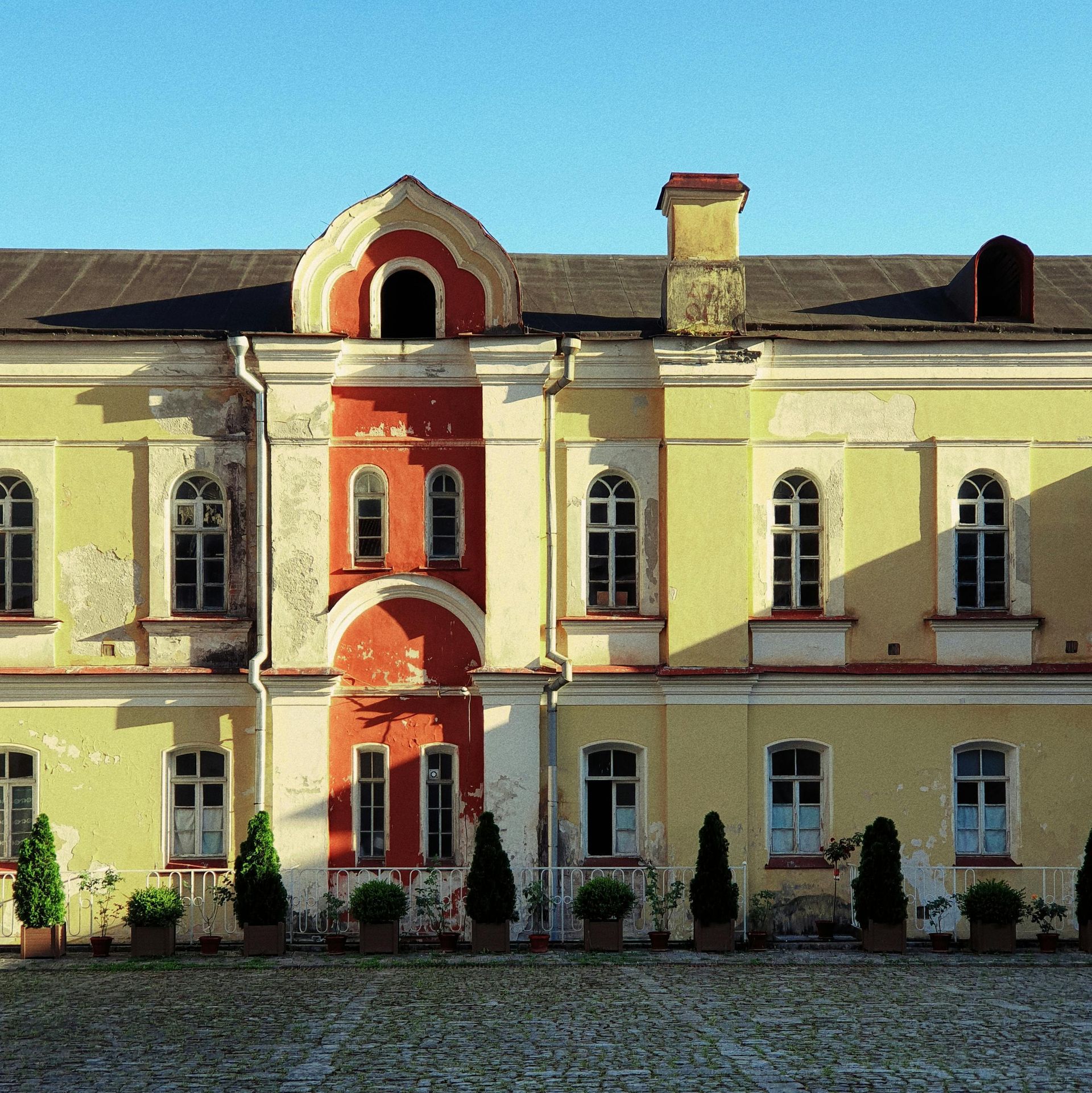 A yellow and red building with many arched windows and potted plants in front of it, set on a stone courtyard.