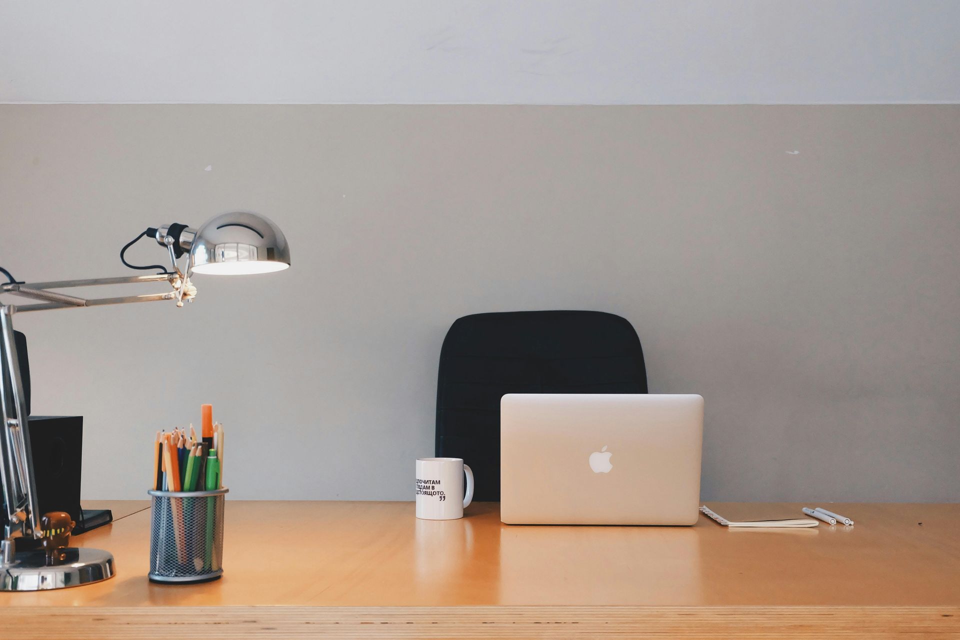 Desk with laptop, mug, pens, lamp, and black office chair in front of a gray wall.