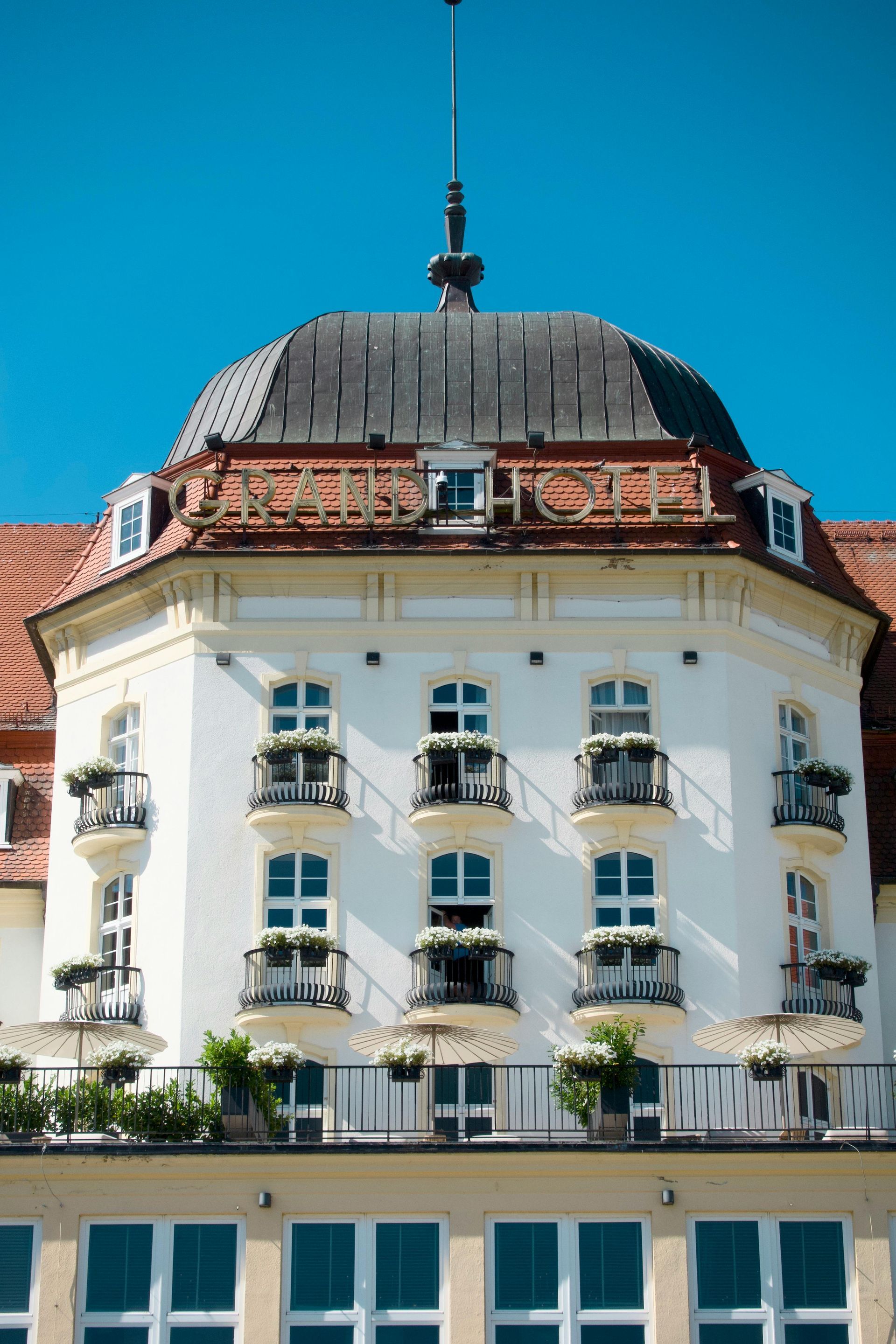 White building with balconies, topped by a dome with a spire. Blue sky backdrop.