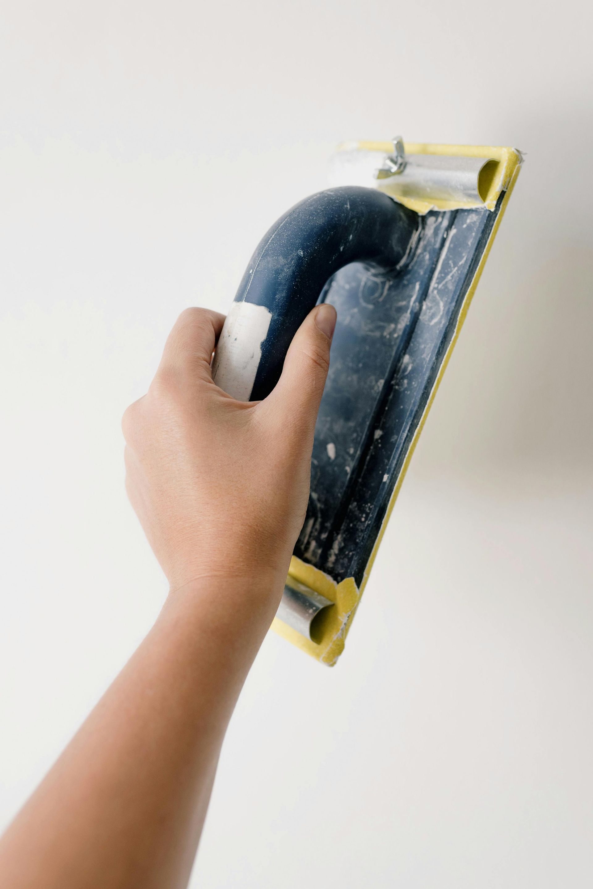 Hand sanding a white wall with a blue and yellow sanding block.