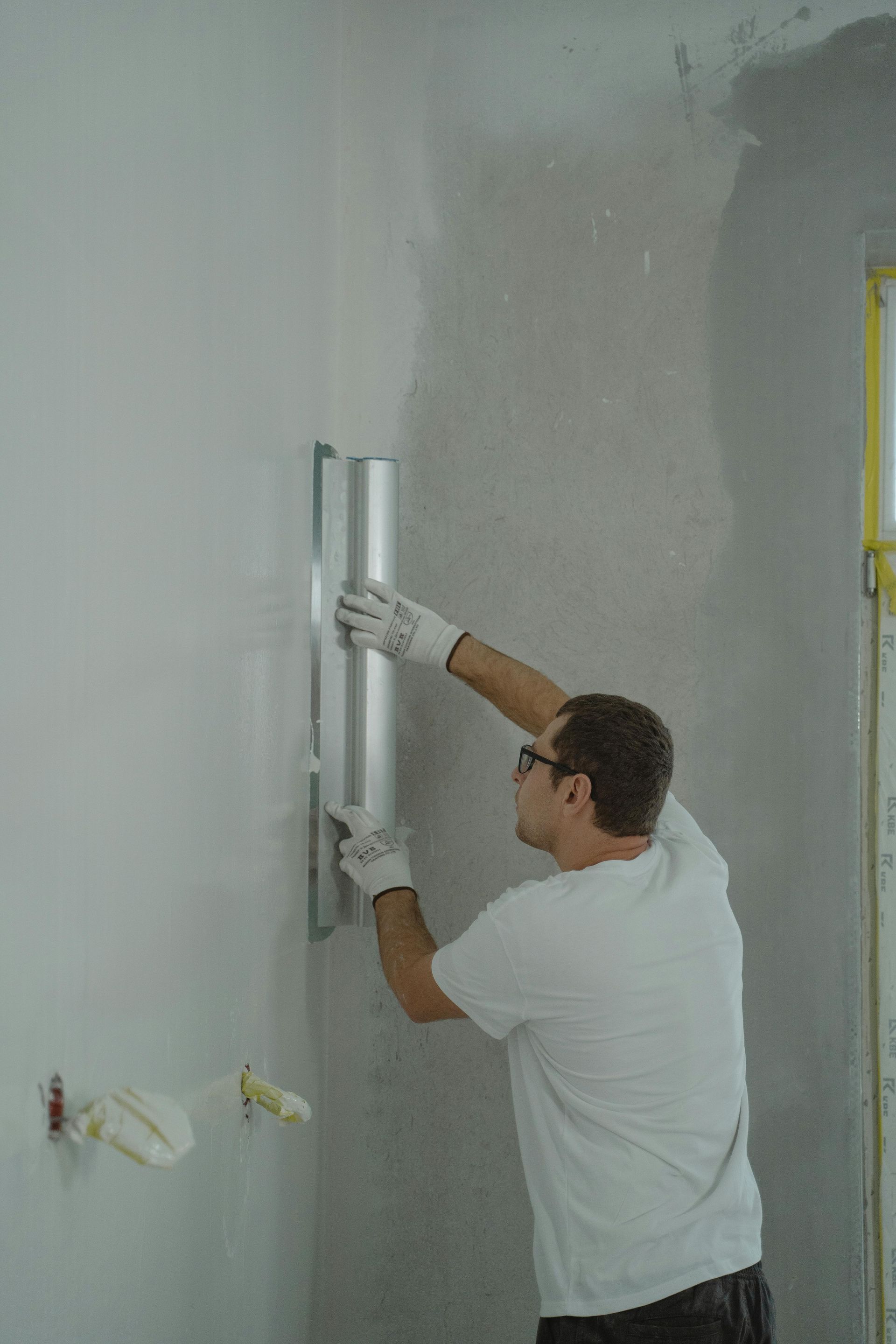 Man smoothing plaster on a wall with a long, metal trowel, wearing gloves, indoors.
