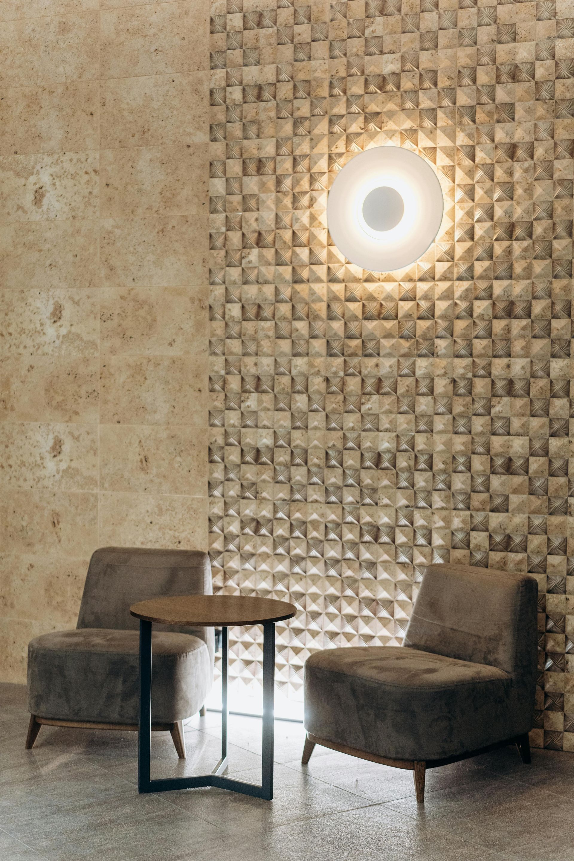Two brown armchairs and a small table in front of a textured wall with a round light fixture.