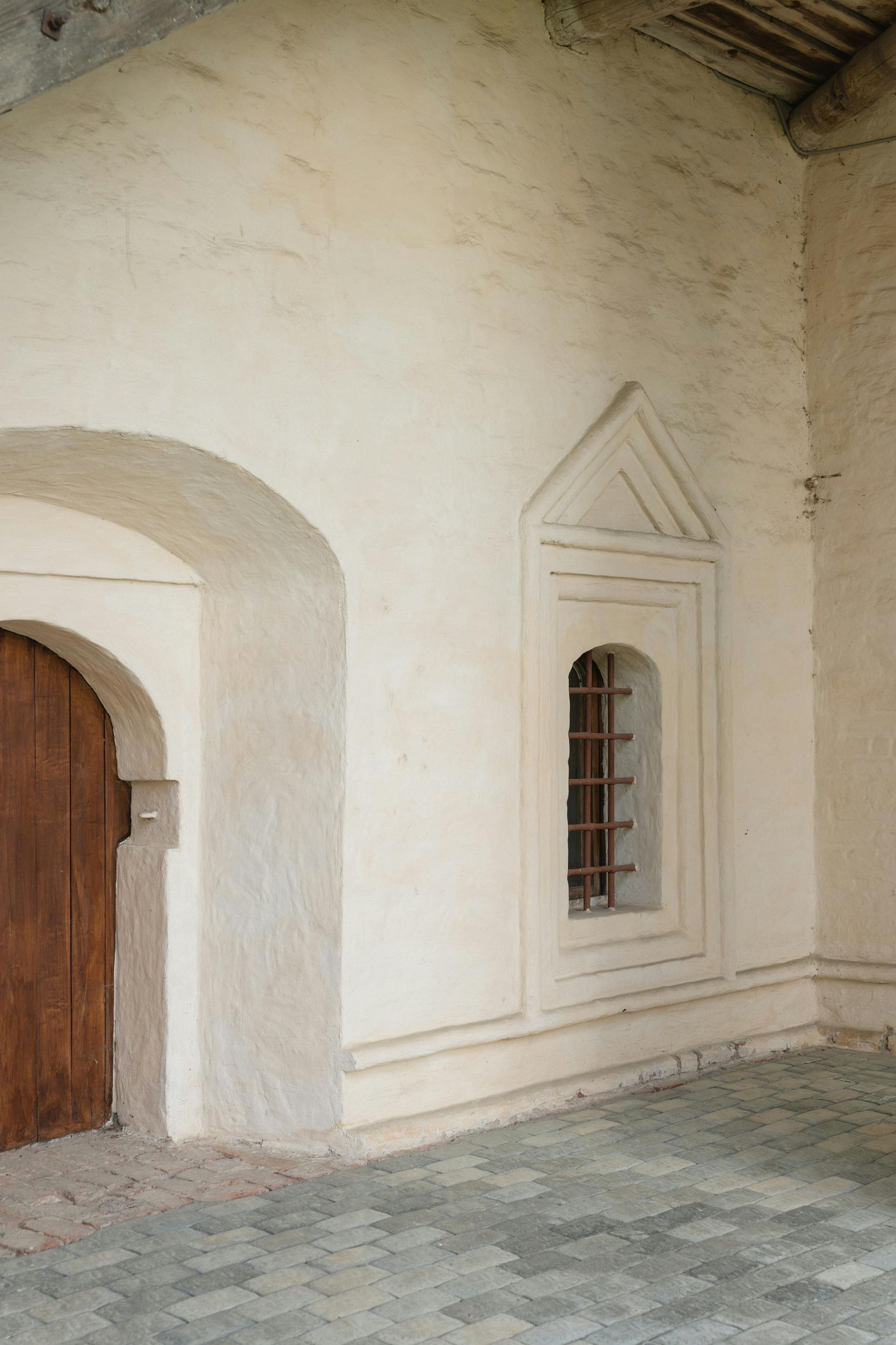 Off-white exterior wall with arched doorway and small barred window. Brown door on the left.