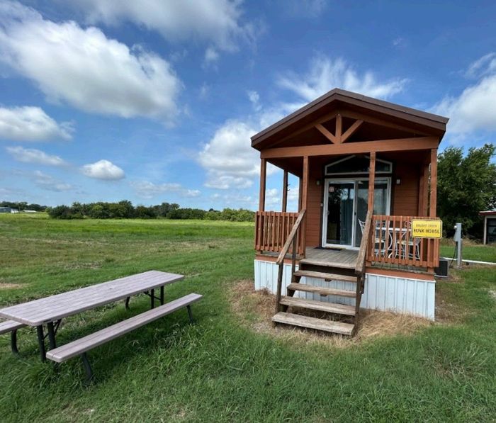 Small cabin with a porch, picnic table, and grassy field under a blue sky.