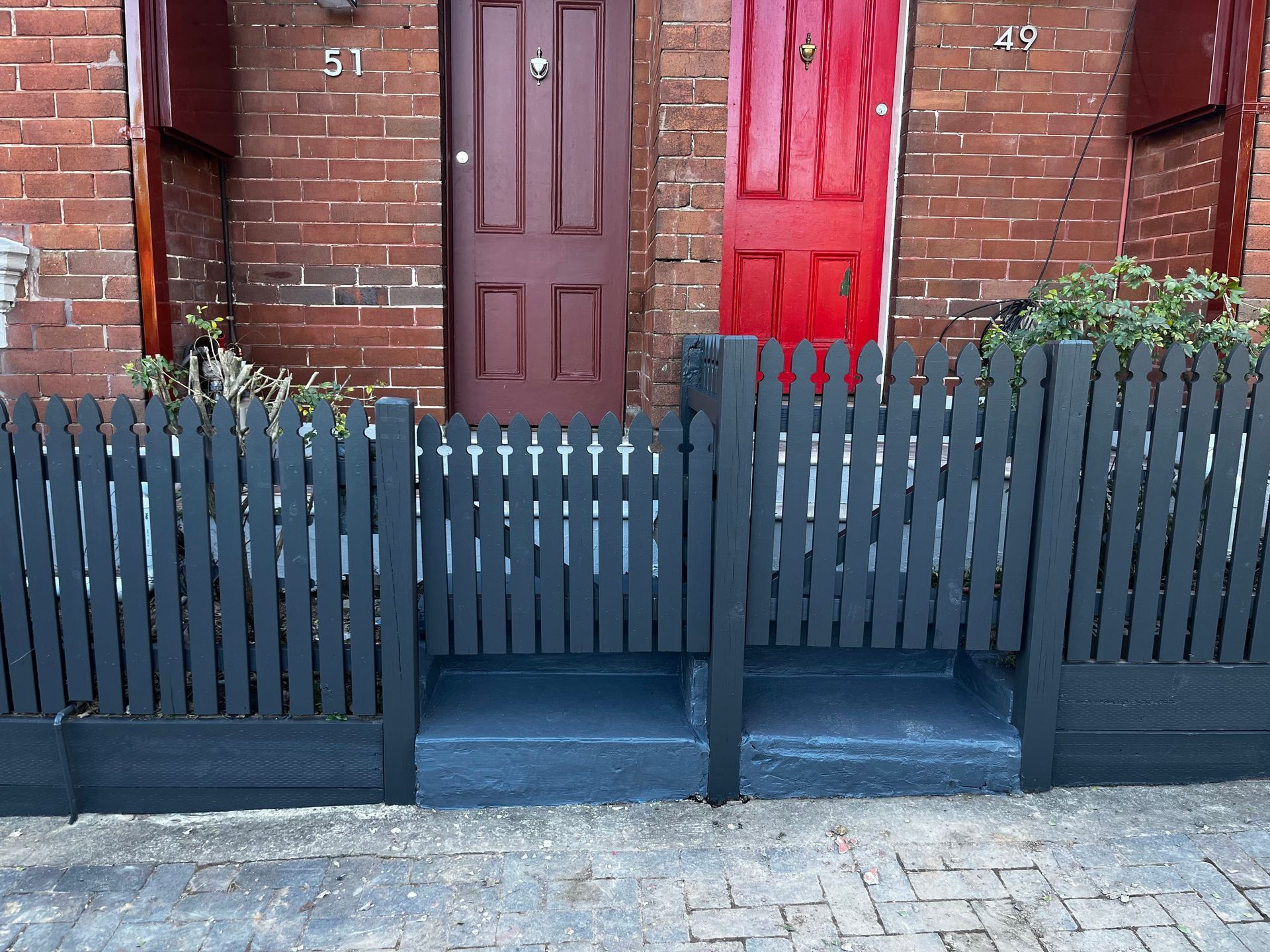 A dark-grey picket fence stands in front of two adjacent brick houses with a dark-maroon door and a bright-red door.