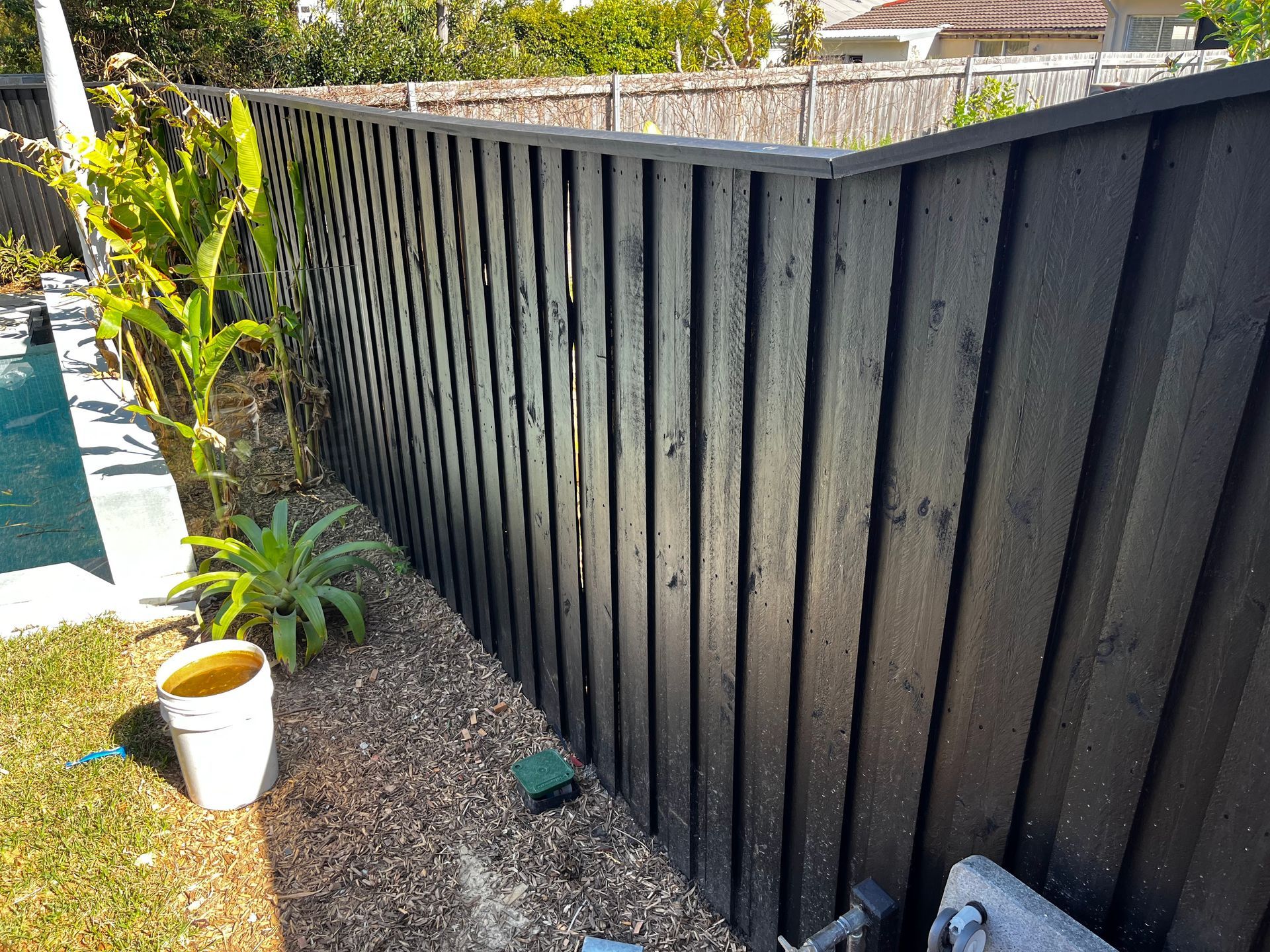 A tall, black wooden vertical board fence stands next to a pool area with a white bucket on the ground.