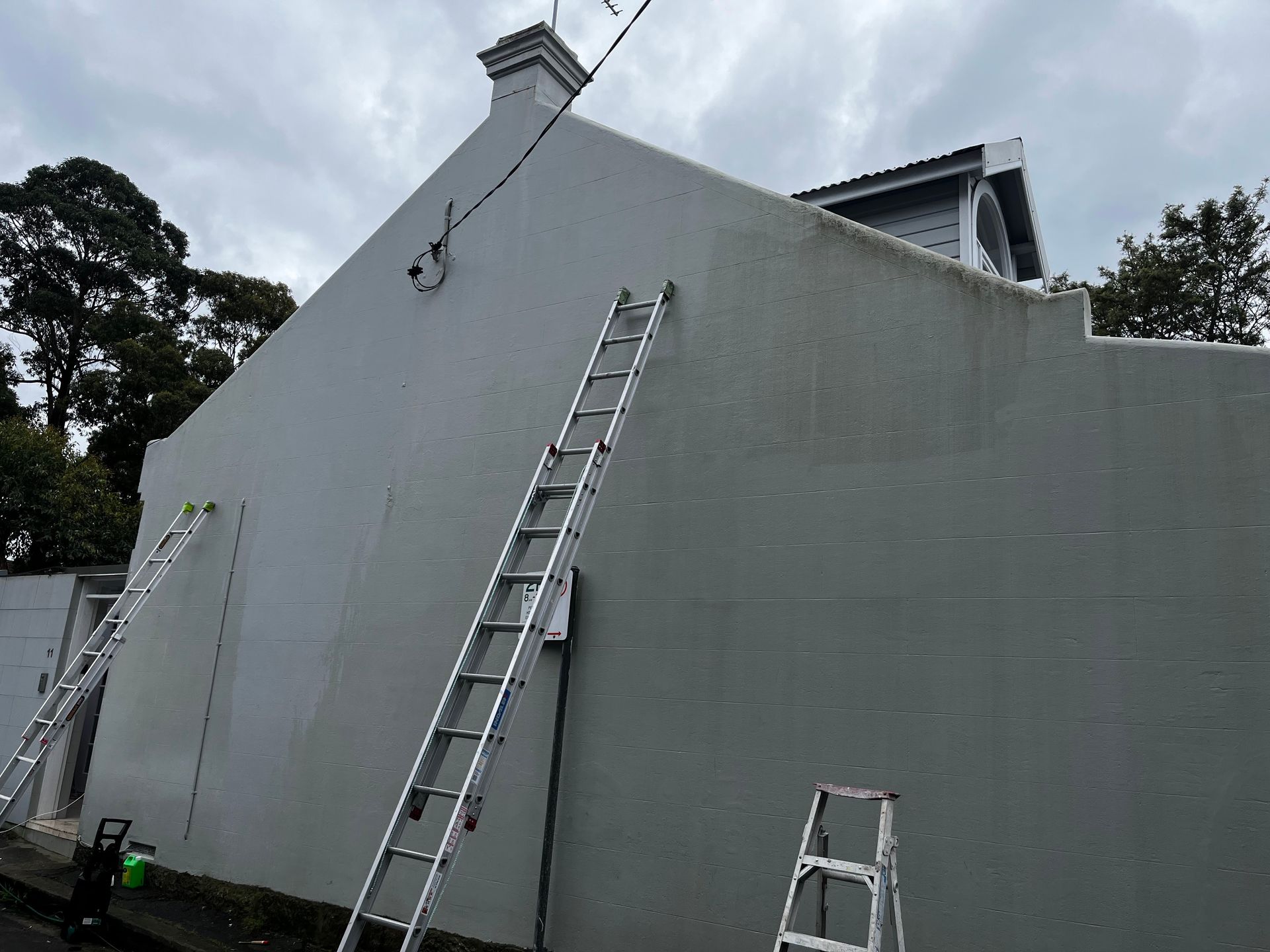 Two metal extension ladders lean against the side of a tall, grey, plastered house wall under a cloudy sky.