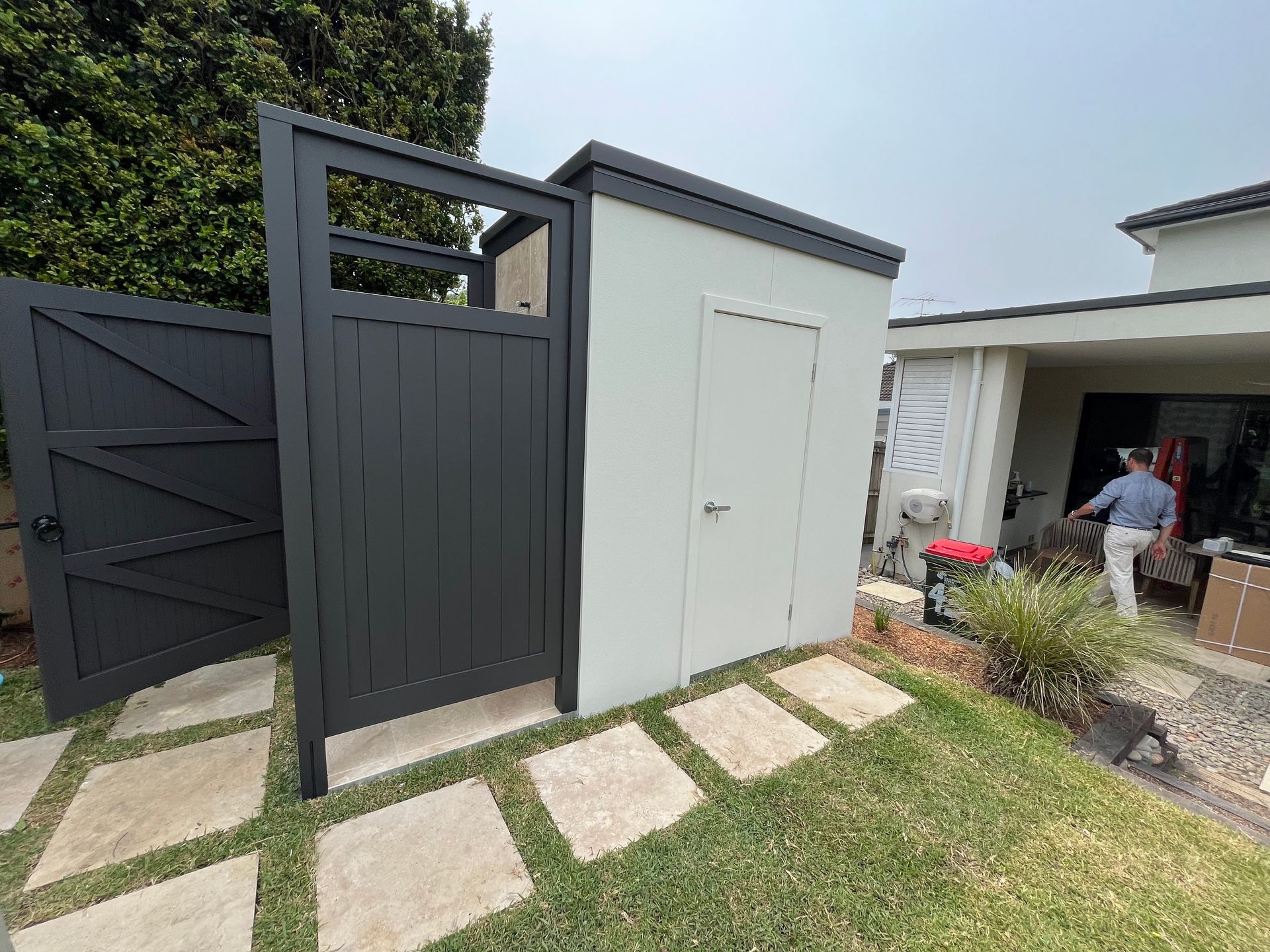 A dark charcoal wooden gate and a light gray storage shed in a backyard with stone pavers leading to a house entrance.