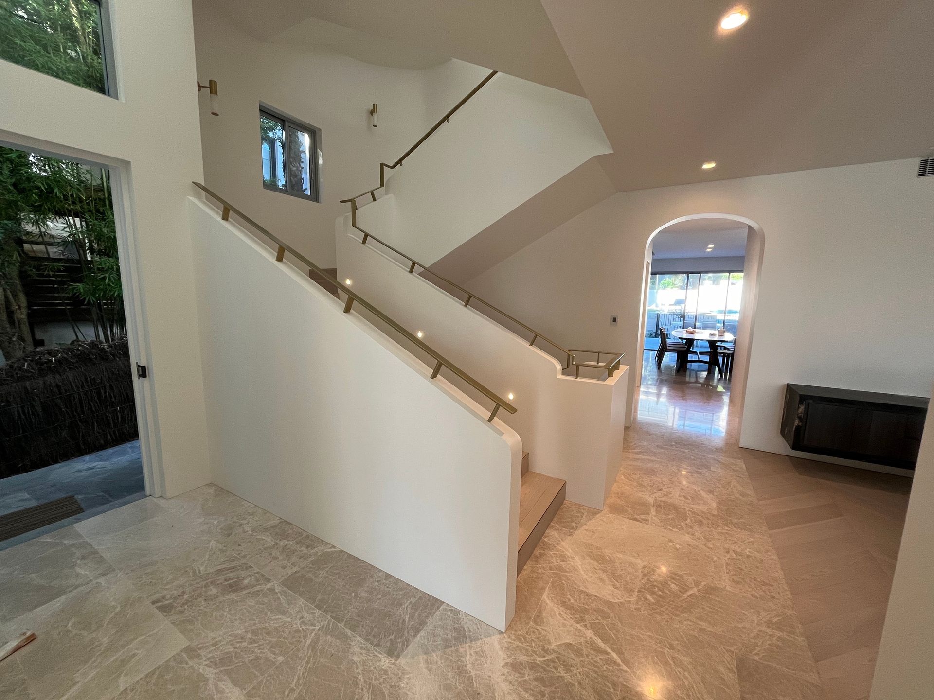 A bright, open foyer featuring a light stone floor, a white staircase with a metal railing, and an arched doorway.