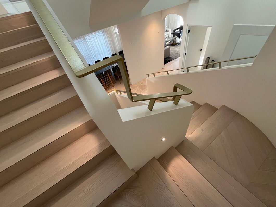 A wooden staircase with beige walls and a modern brass railing, viewed from the top landing looking down.