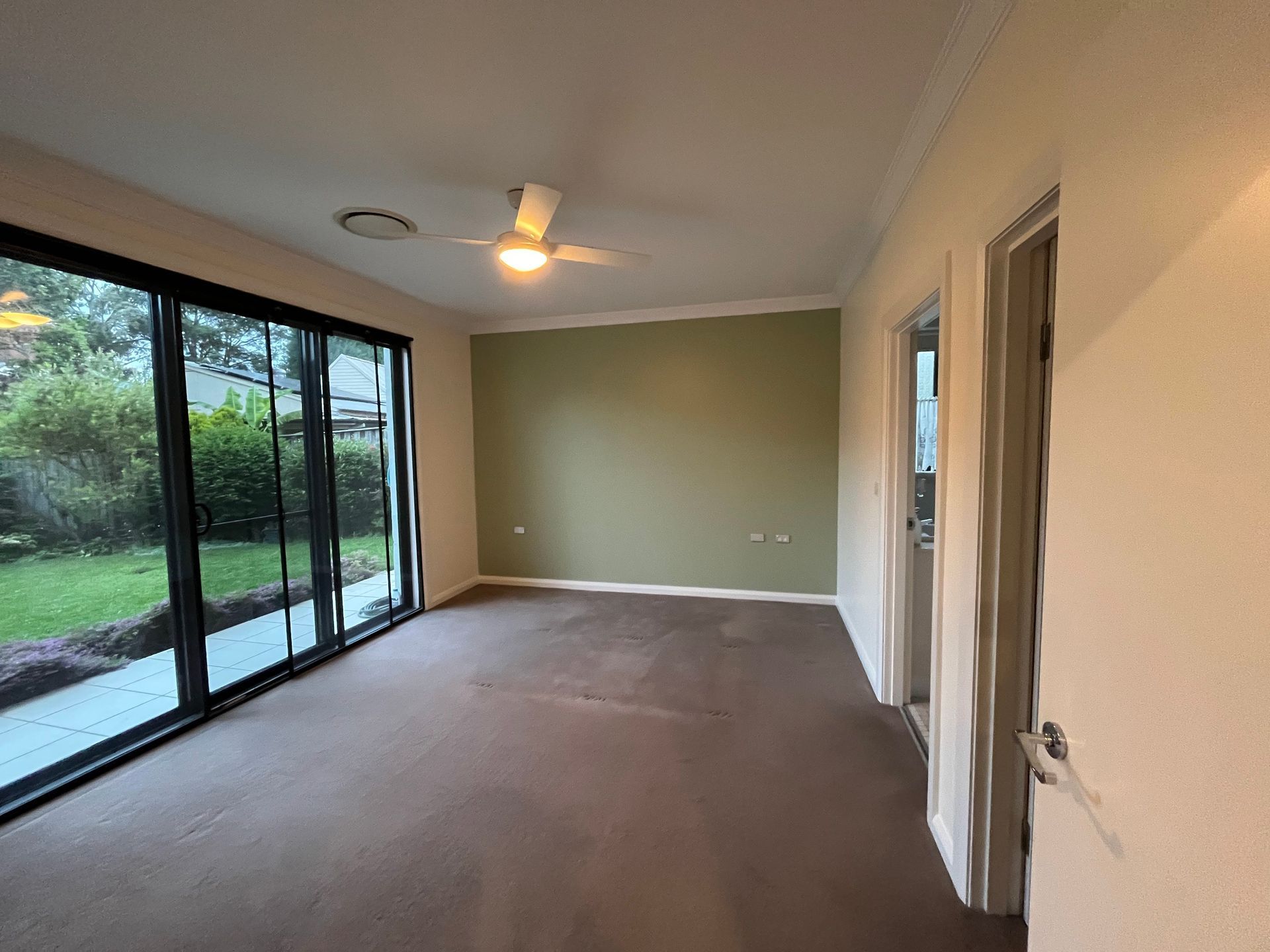 An empty room with tan carpet, a green accent wall, a ceiling fan, and sliding glass doors looking out to a garden.