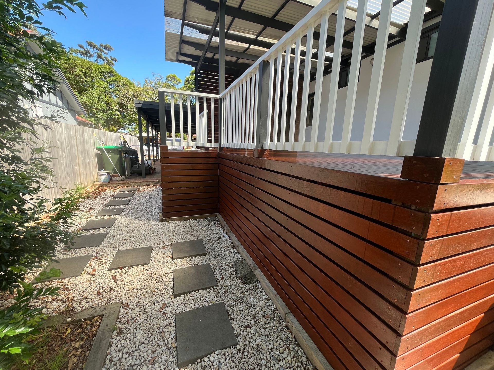 A side view of a raised wooden deck with white railings, set beside a stone path with stepping stones in a backyard.