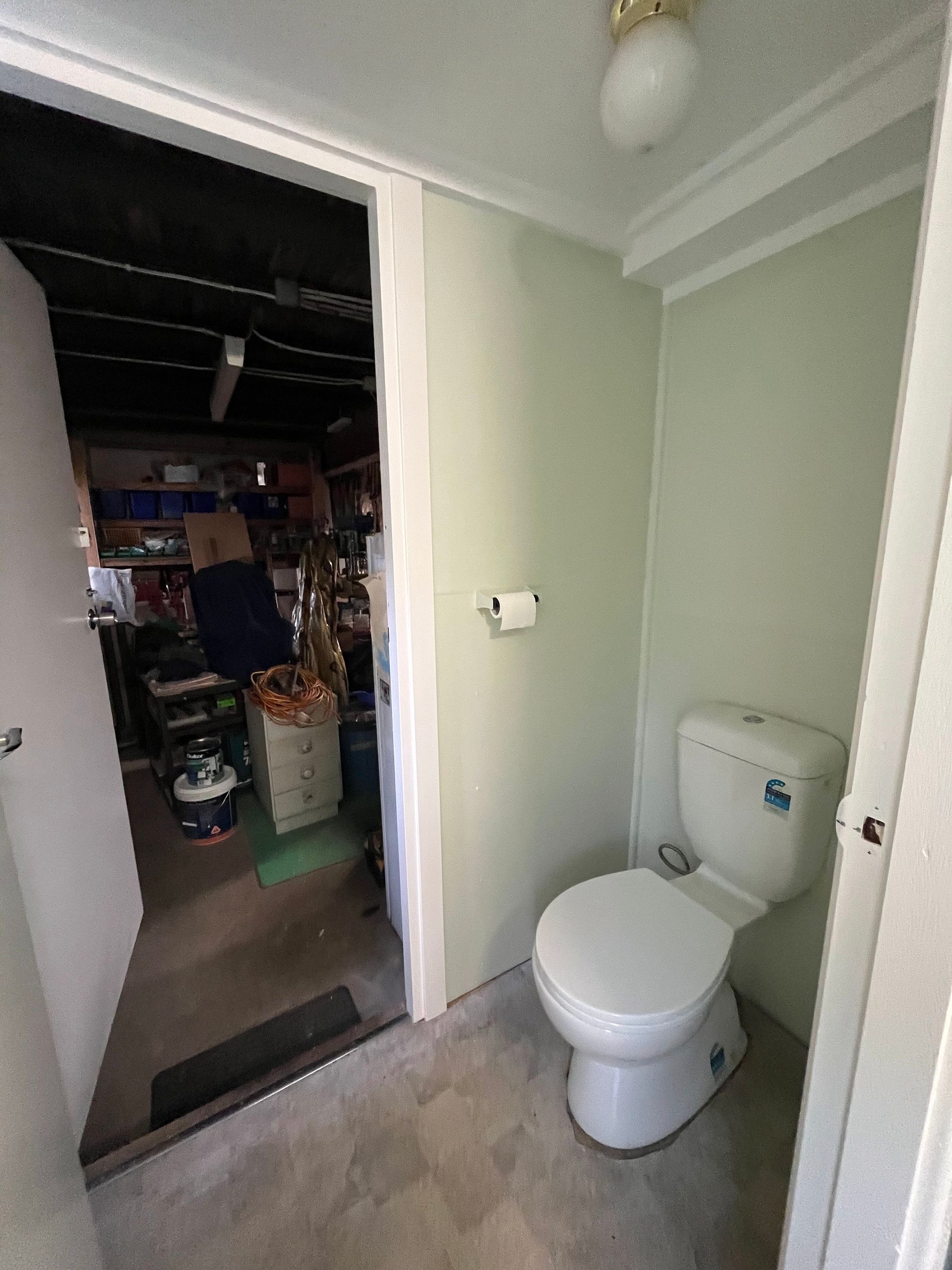A white toilet in a light green room, with an open doorway leading into a cluttered, dark storage area.
