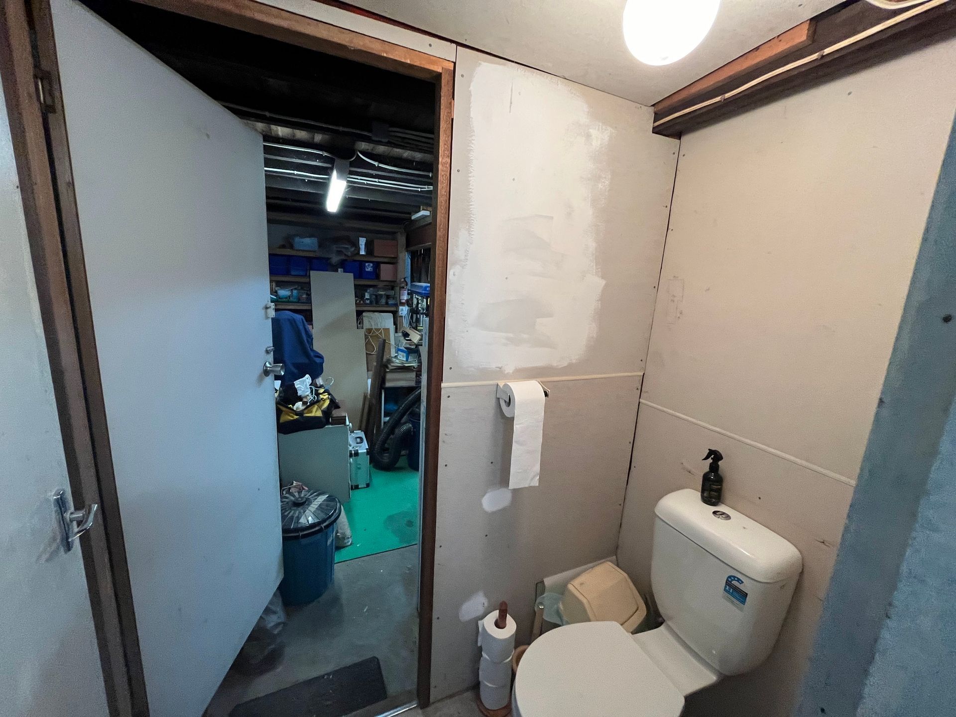 A toilet room with unpainted drywall walls, a white toilet, and a doorway leading to a cluttered workspace.