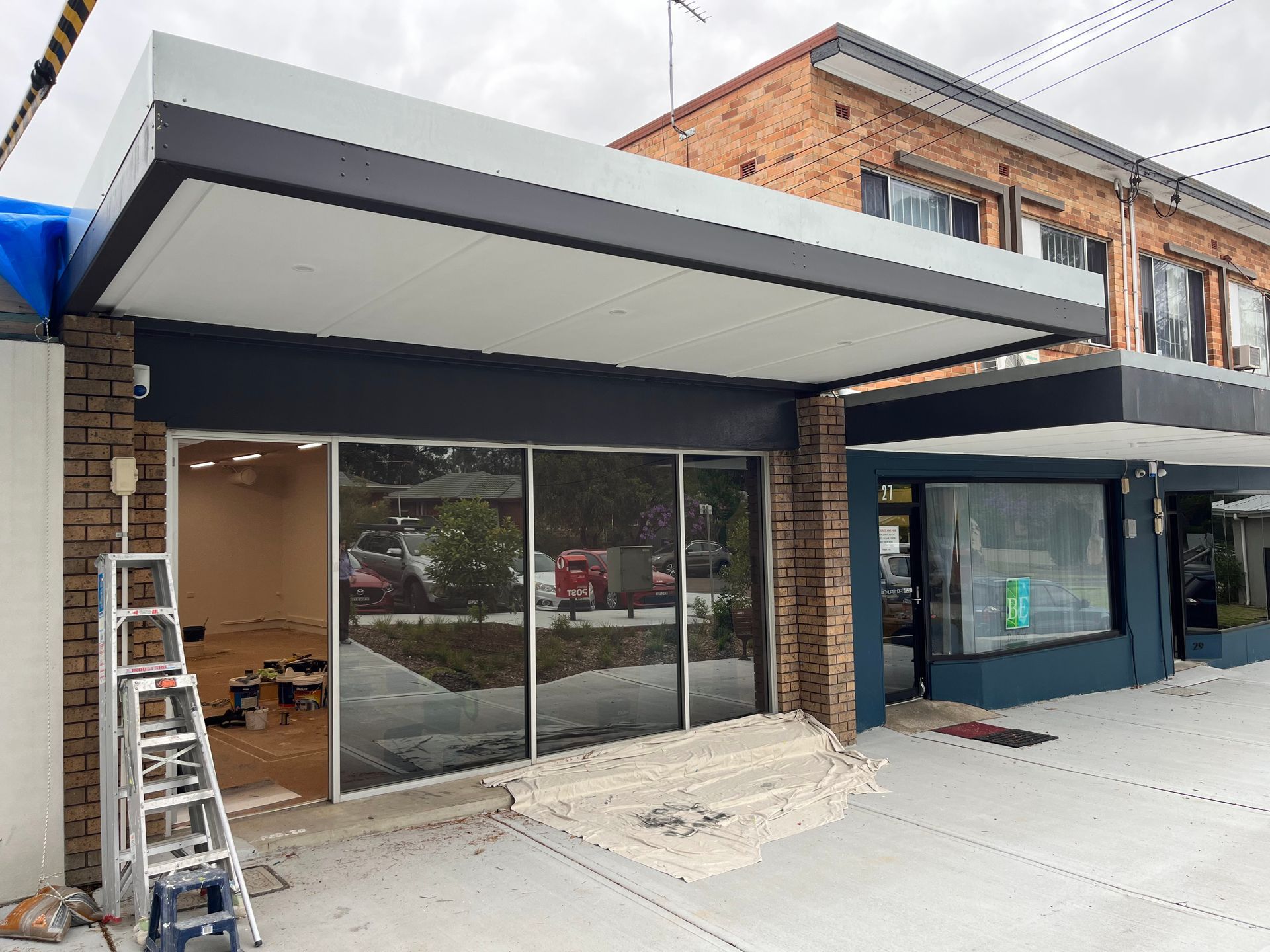 A storefront with brick pillars, a modern white and dark gray awning, and large glass windows, with a ladder outside.