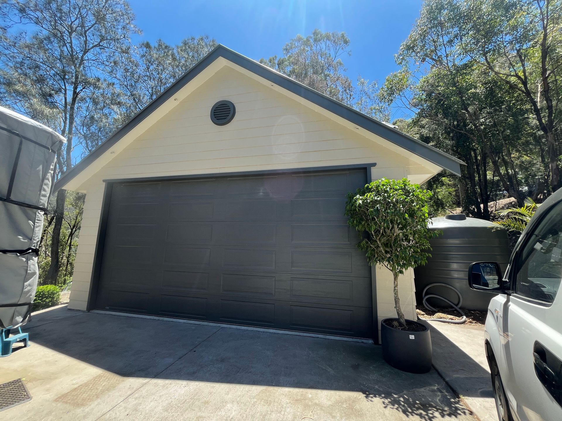 A detached garage with a dark sectional door, cream-colored siding, and a small potted tree against a sunny backdrop.