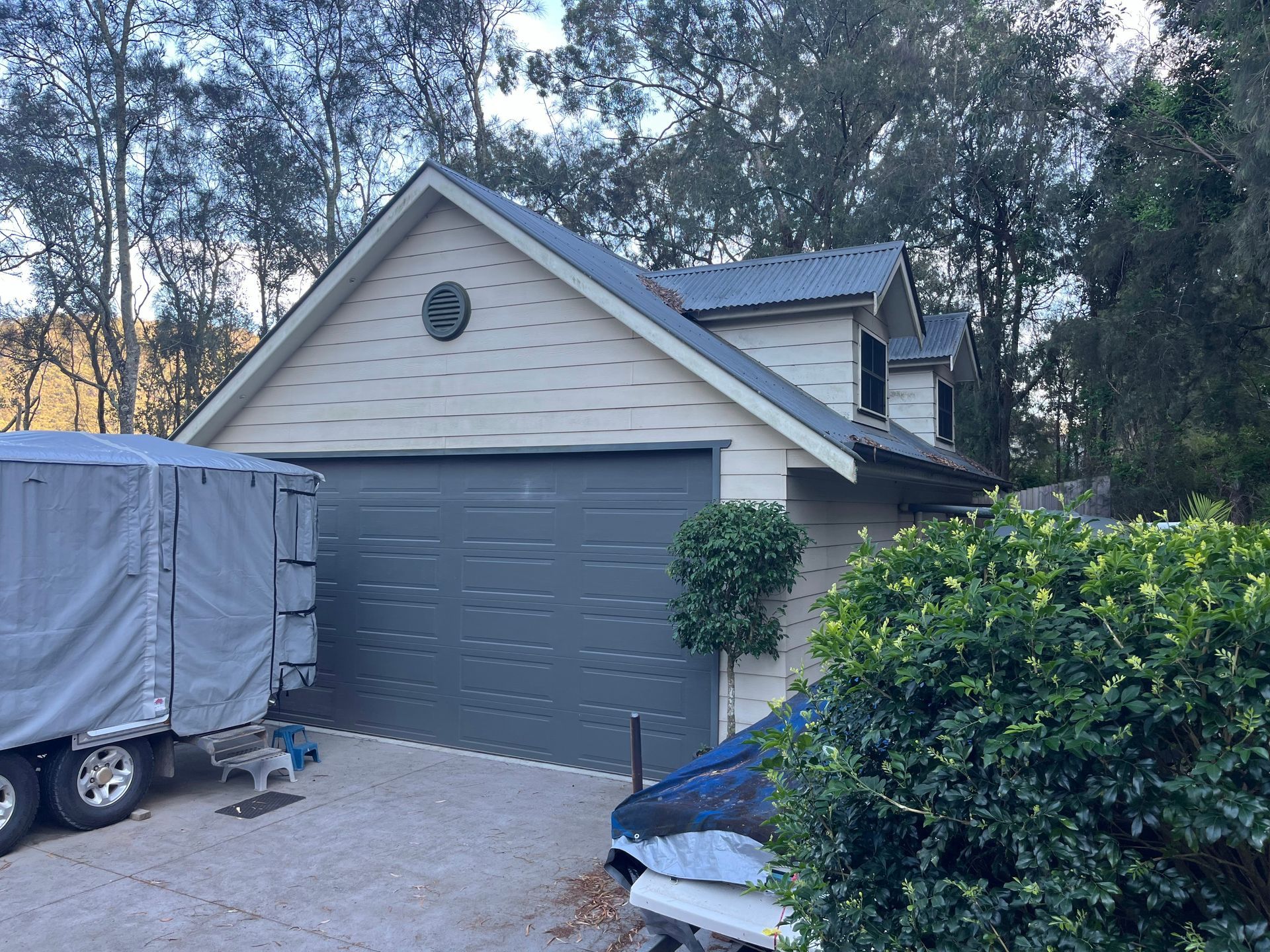 A beige detached garage with a dark gray door and roof, parked next to a large covered trailer and a bush.