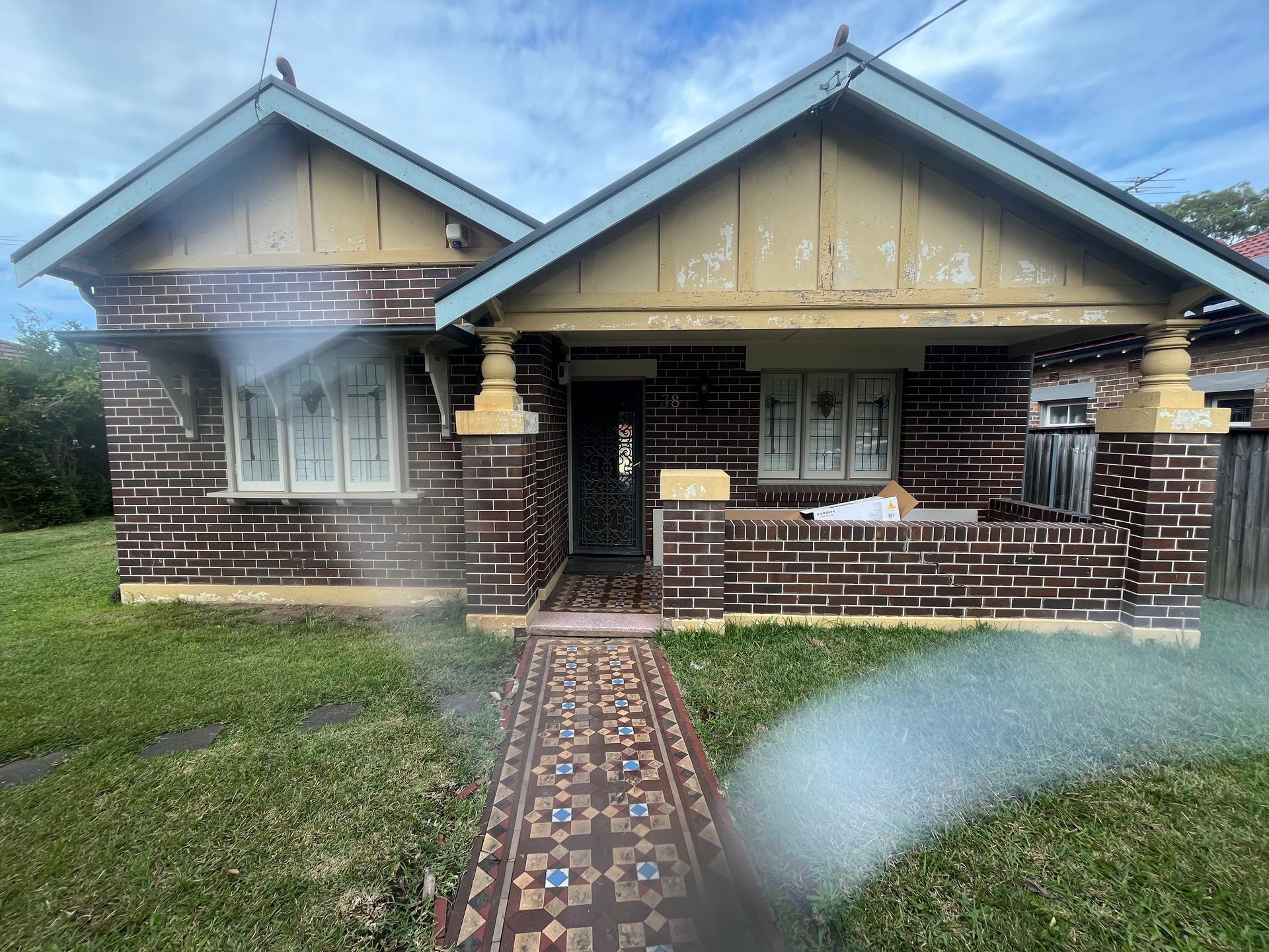 A house with brown brick walls, a tiled walkway, and two gabled roofs, with light haze over the front lawn.