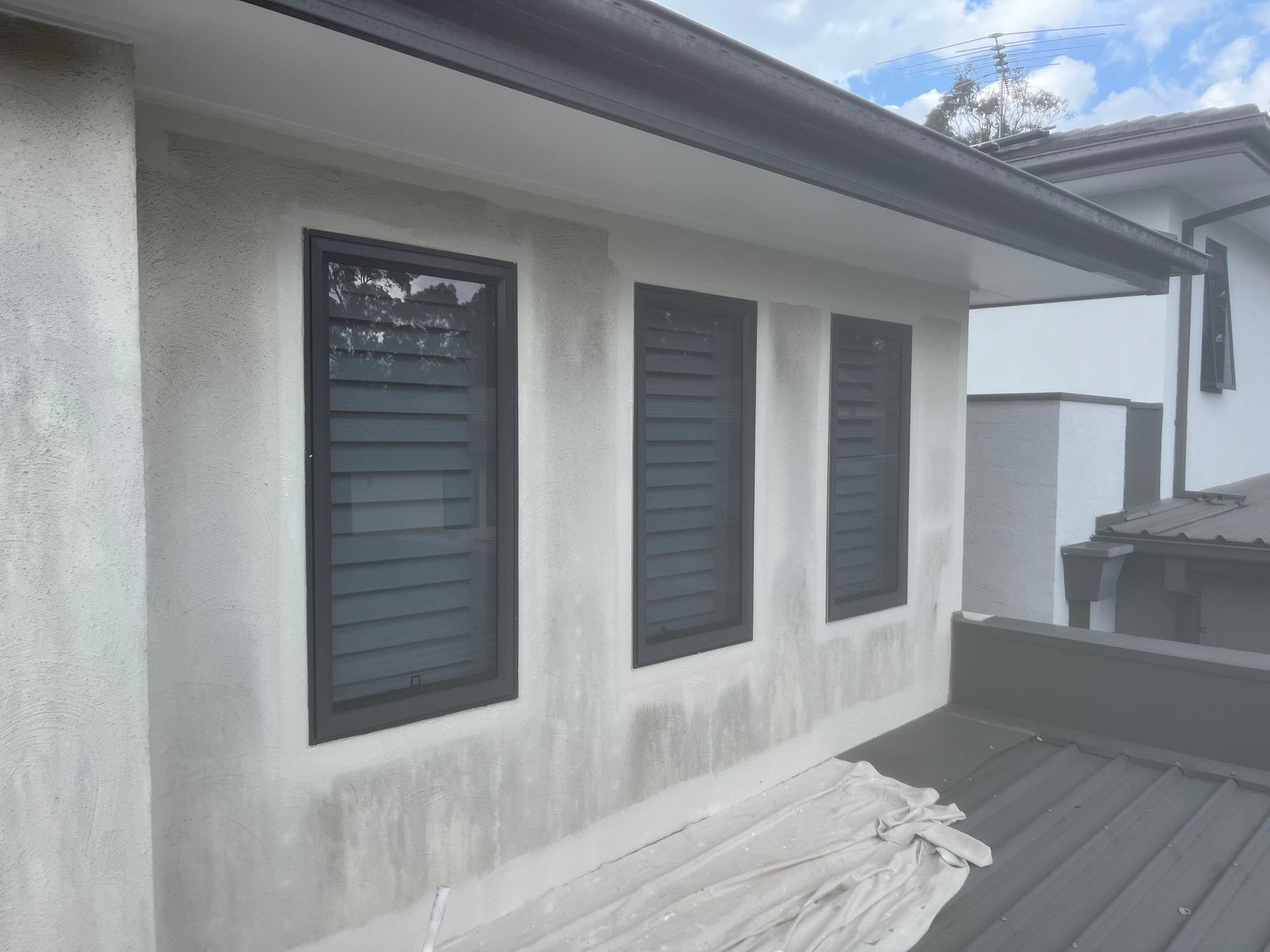 An exterior view of a building wall with three tall, dark-framed louvered windows above a dark rooftop.