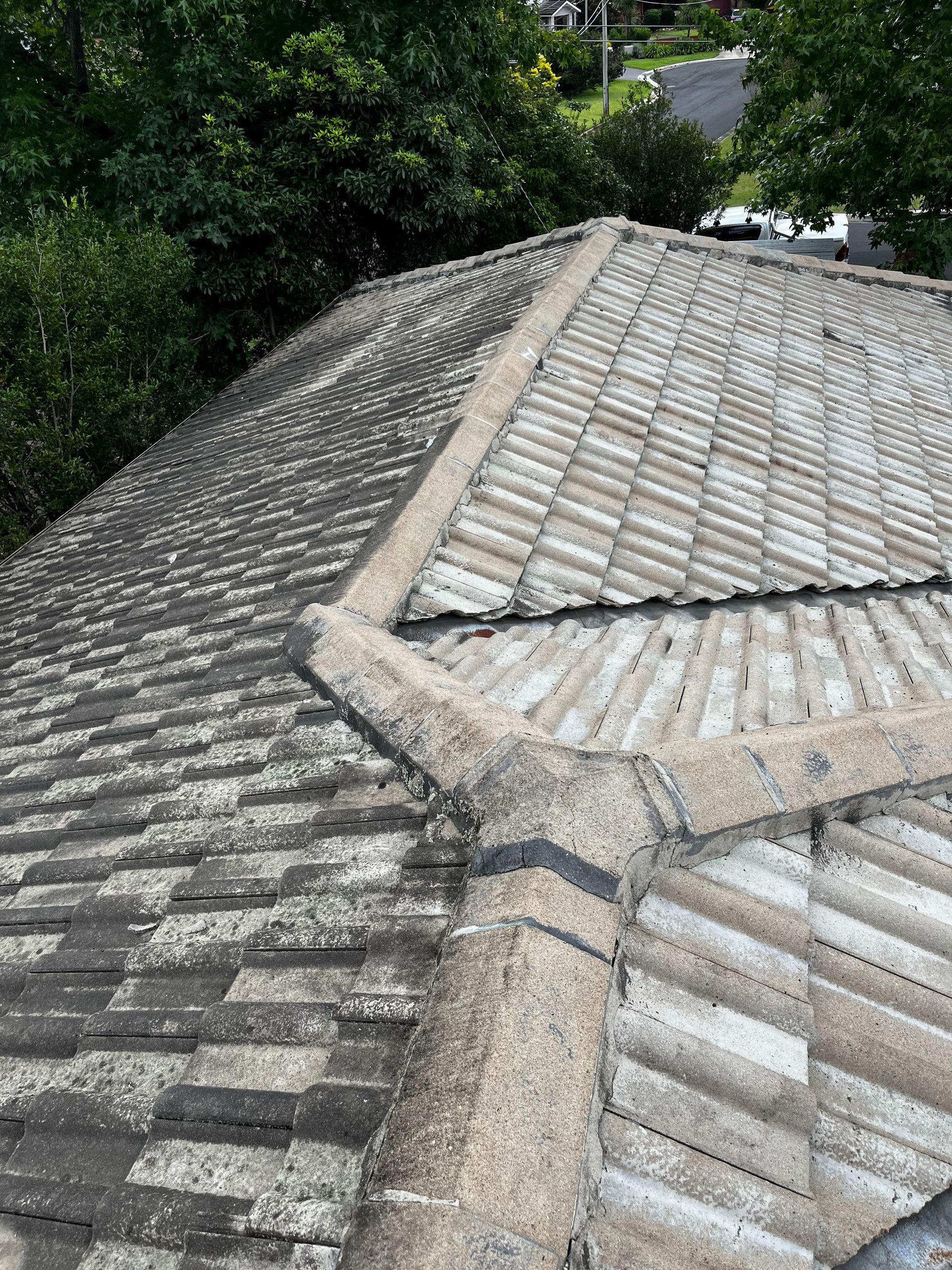 A top-down view of a weathered, gray-tiled roof with a ridge line and hip sections, surrounded by trees and a street.