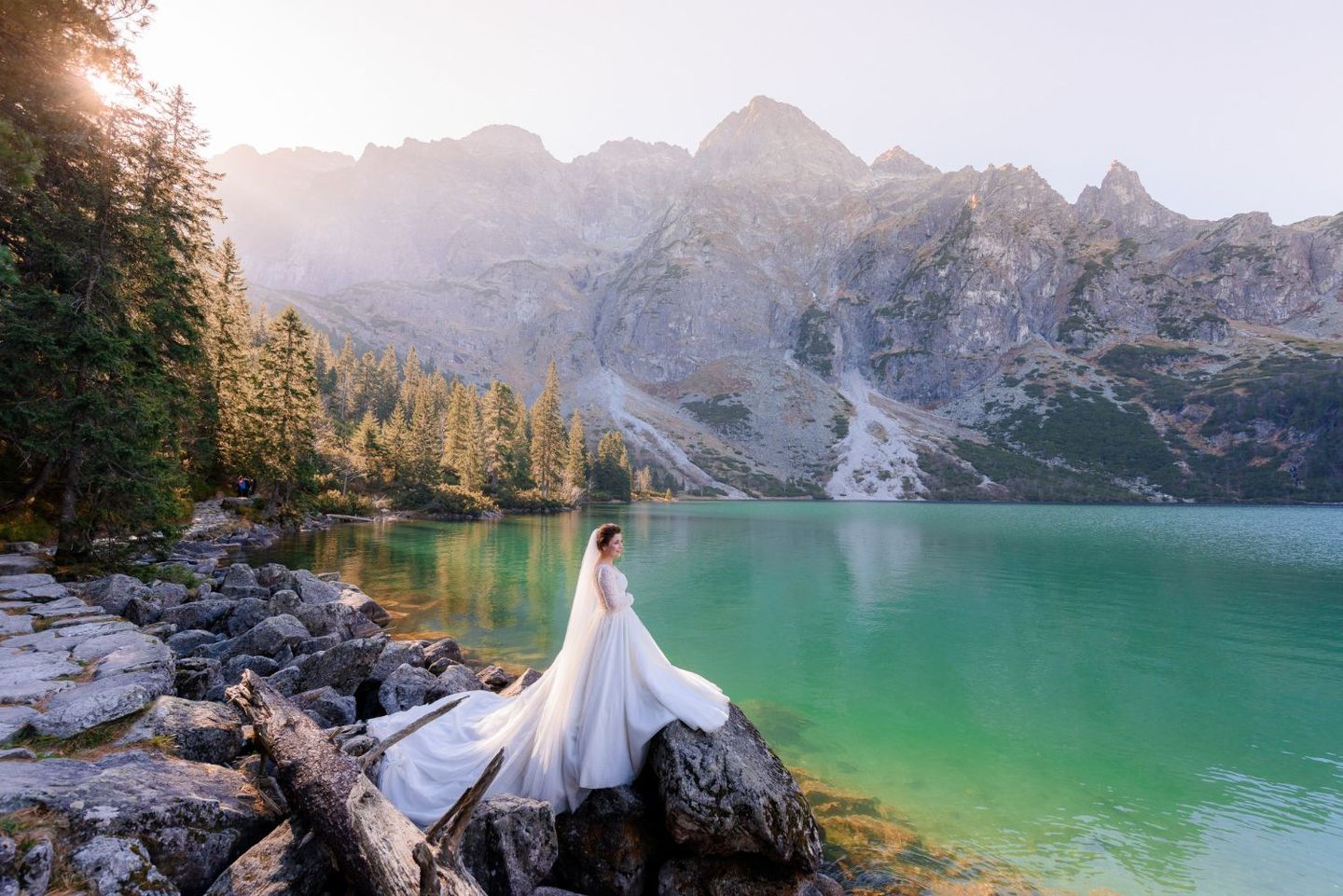 A bride in a long white gown sits on a rocky shoreline by a teal mountain lake under a bright, hazy sky.