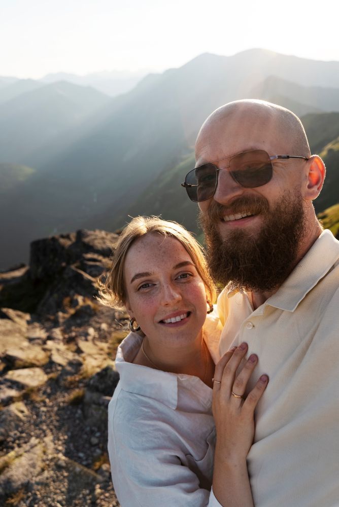 Couple smiling in mountains. Man with beard and sunglasses, woman in white shirt. Sunny, outdoor setting.