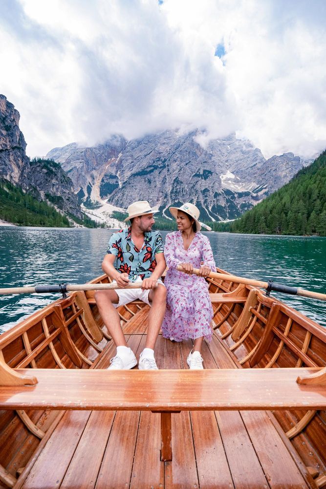 Couple rowing a wooden boat on a lake, mountains in background. Cloudy sky.