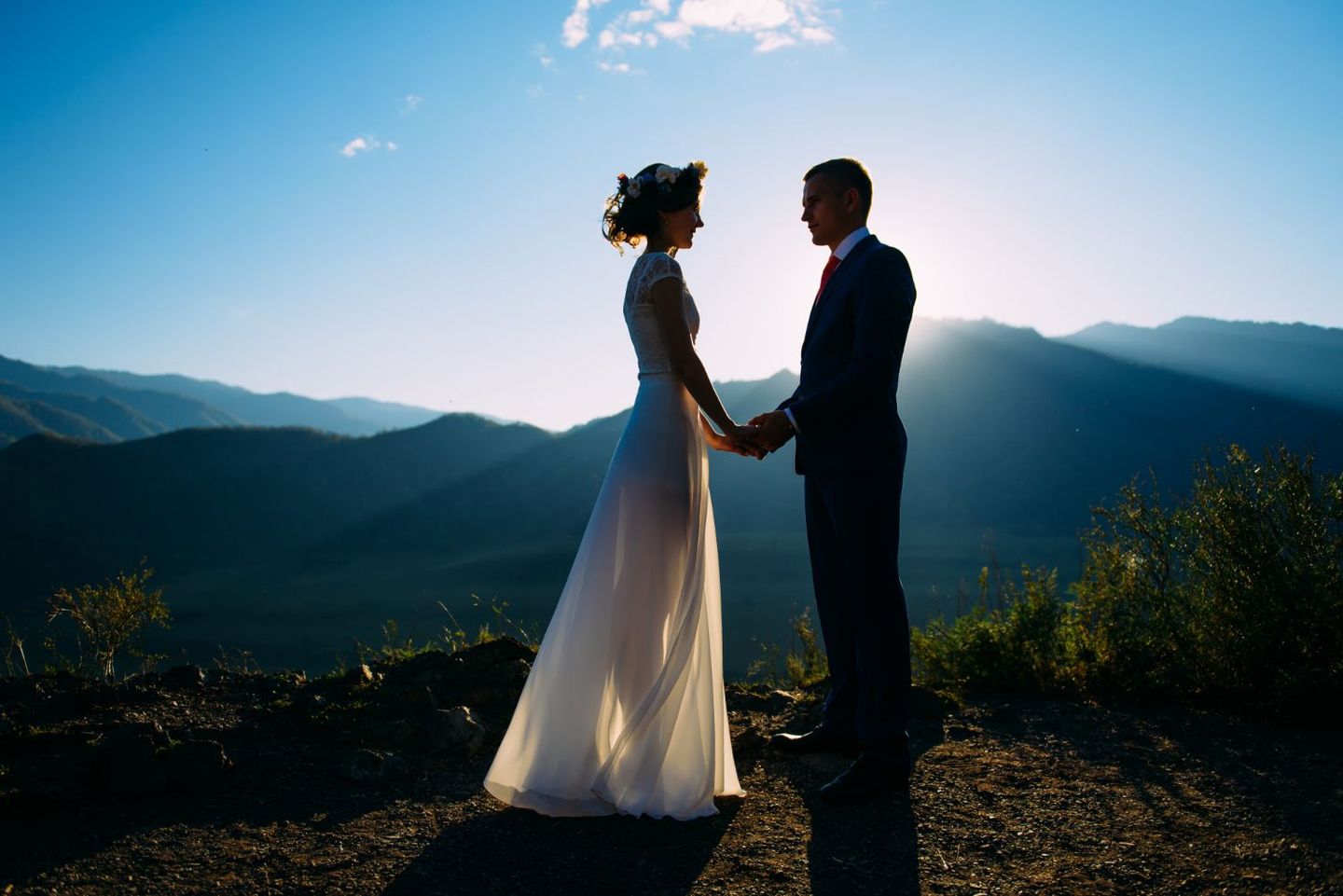 A bride and groom hold hands in silhouette against a bright mountain sunset.