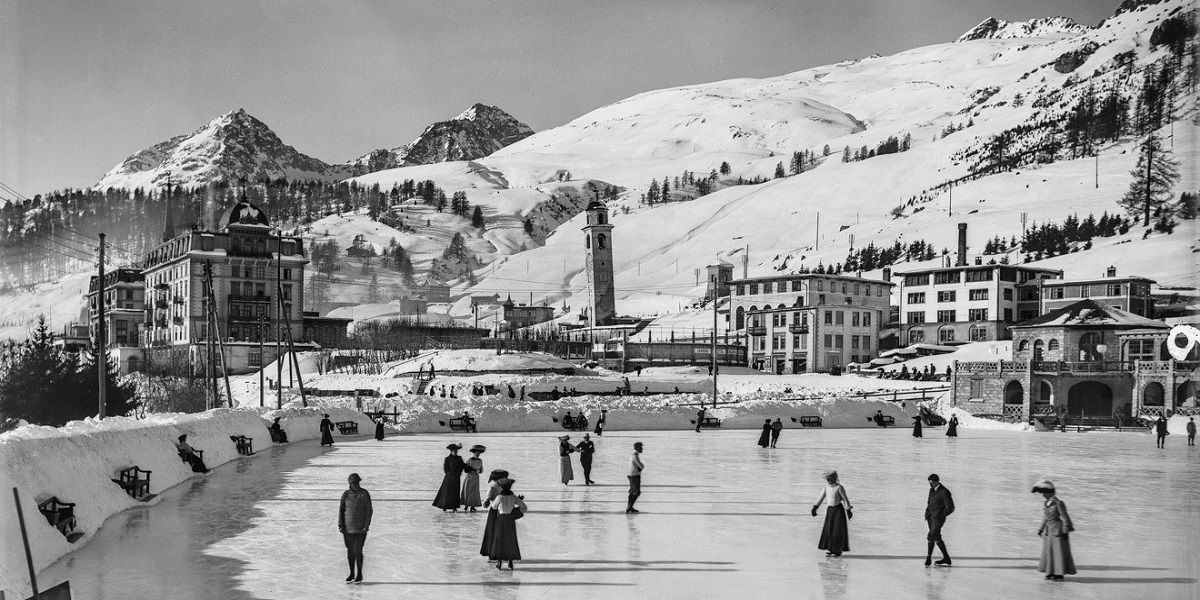 People ice skate on a frozen lake with snowy mountains and buildings in the background. Black and white.