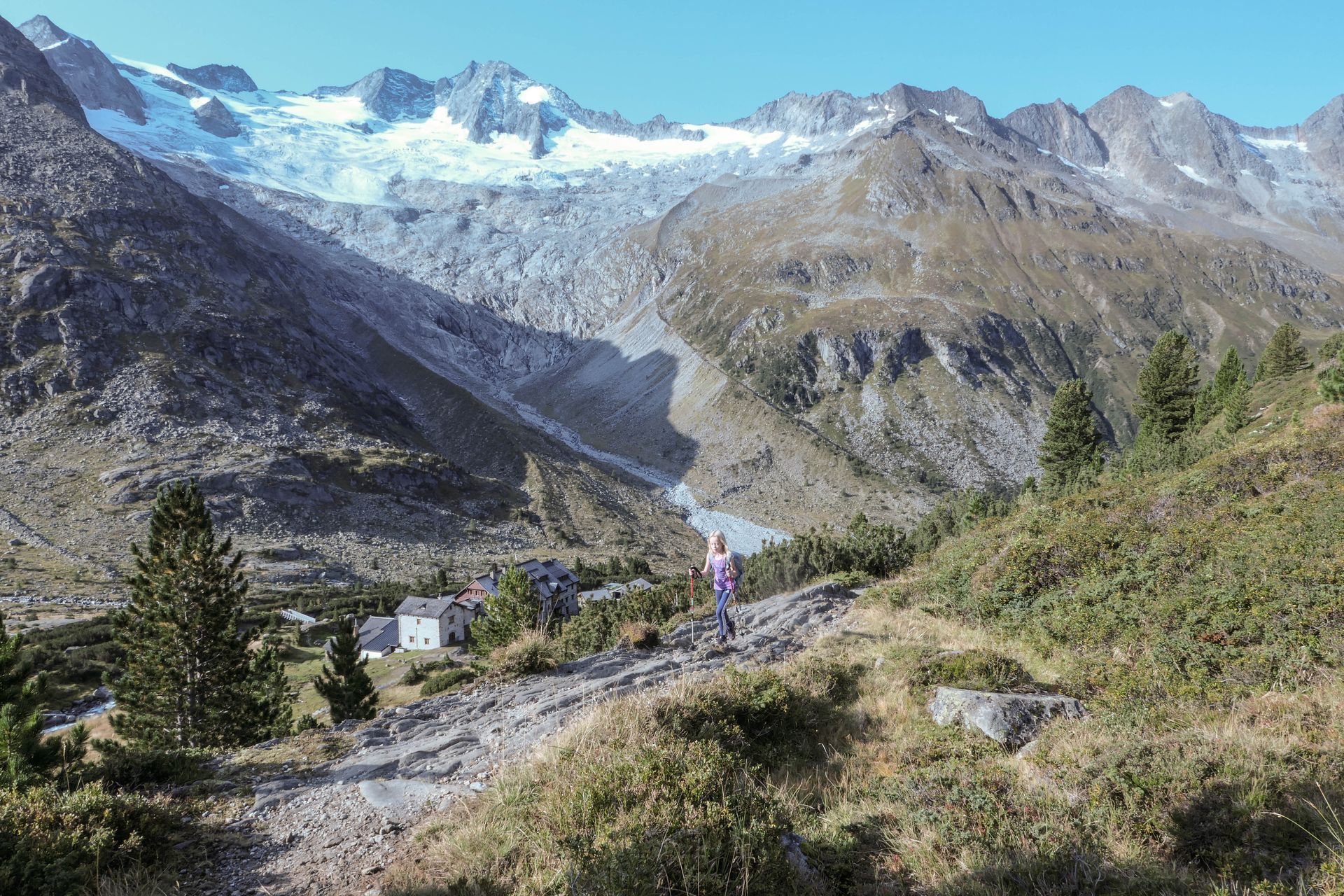Mountain trail leading to a valley with buildings, a glacier, and peaks under a blue sky.