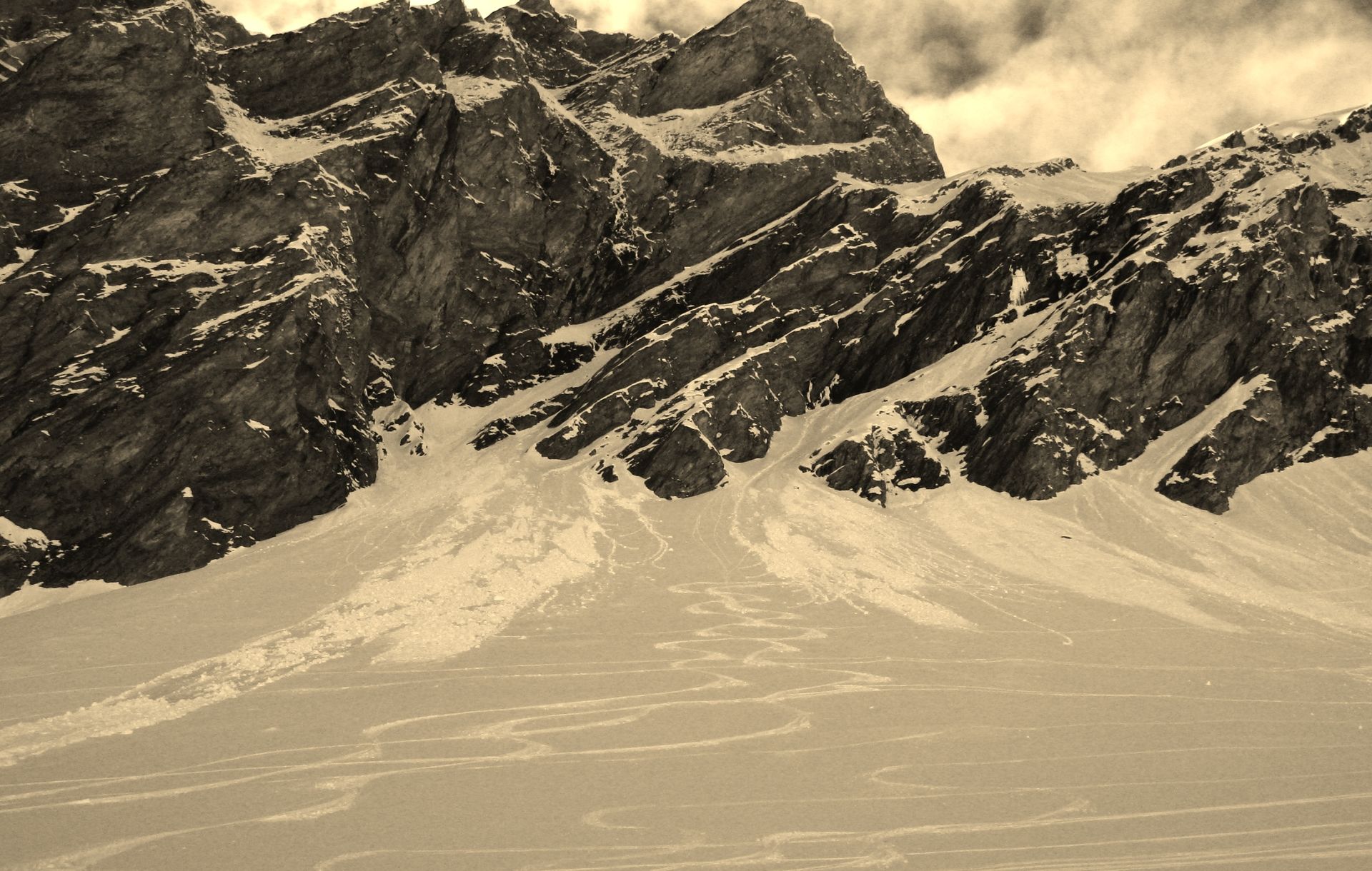 Snow-covered mountain range with dark, rocky slopes. Cloudy sky above.