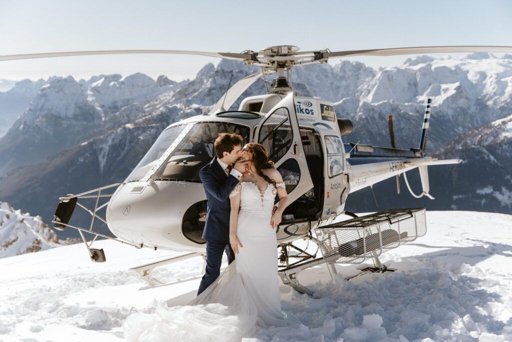 Couple kissing in wedding attire next to a helicopter on a snowy mountain peak.