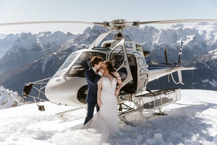 Couple kissing in wedding attire next to a helicopter on a snowy mountain peak.