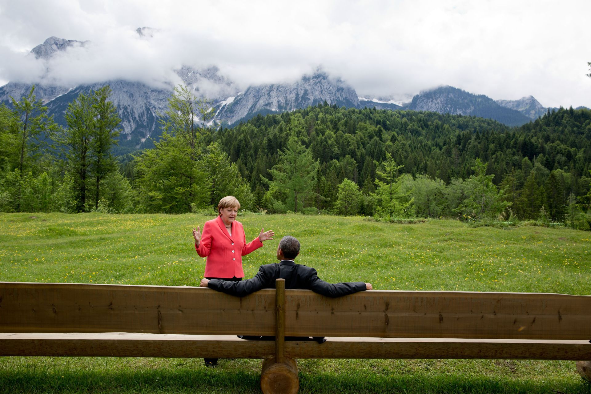 Two people conversing outdoors, seated on a wooden bench, with mountains and forest in the background.