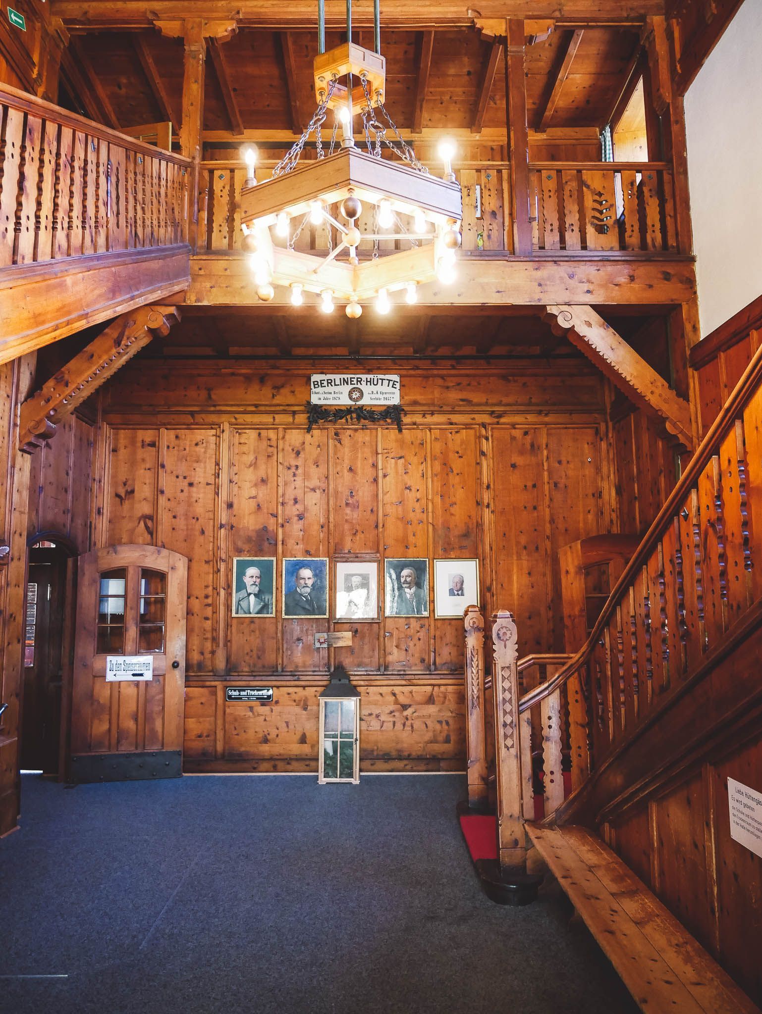 Wooden interior with staircase, chandelier, portraits, and a lantern.