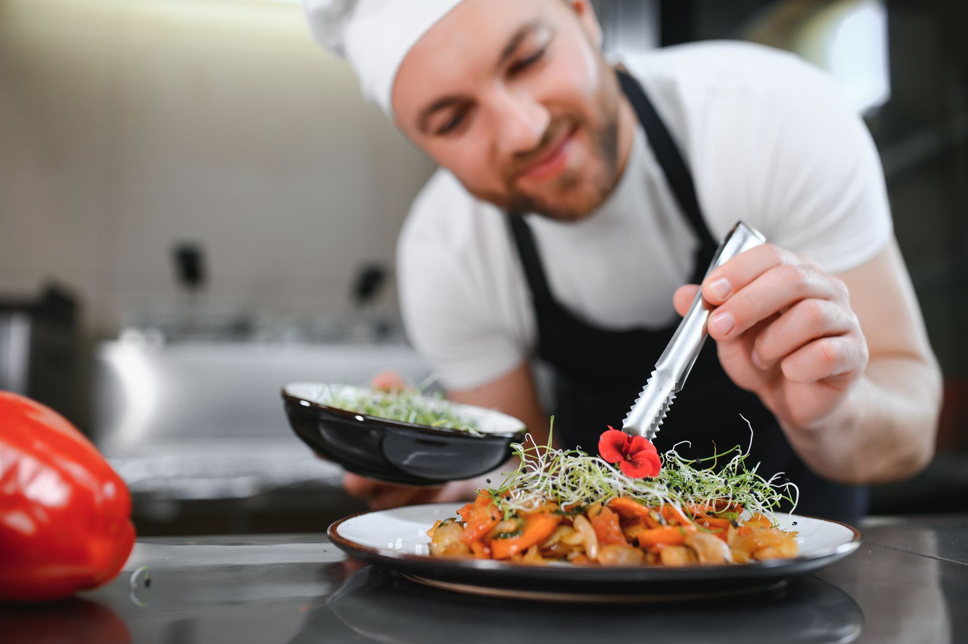 Chef garnishes a dish with microgreens and an edible flower in a kitchen.