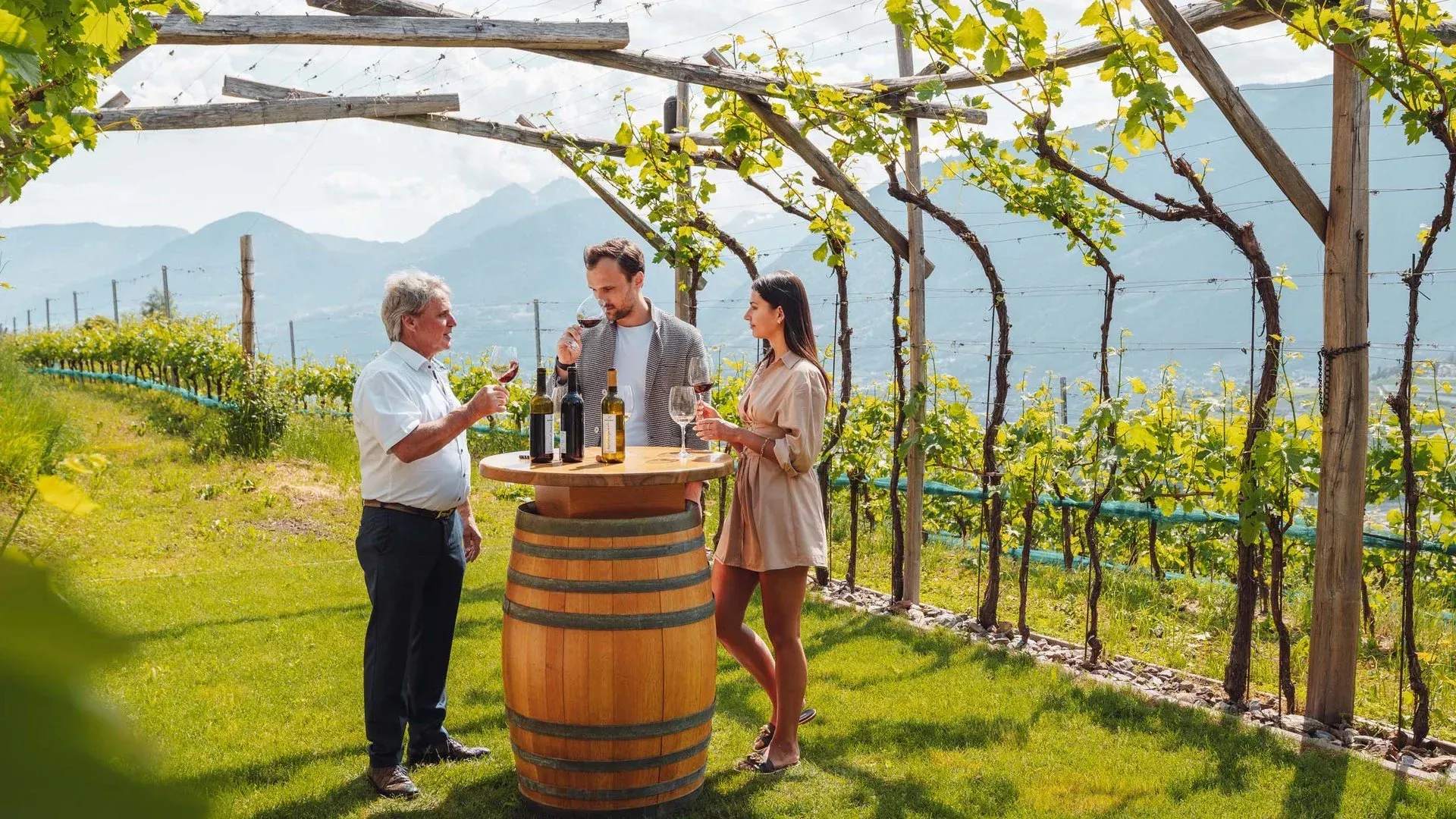 People tasting wine at a vineyard. Man pours wine, standing near a barrel table, with mountain backdrop.