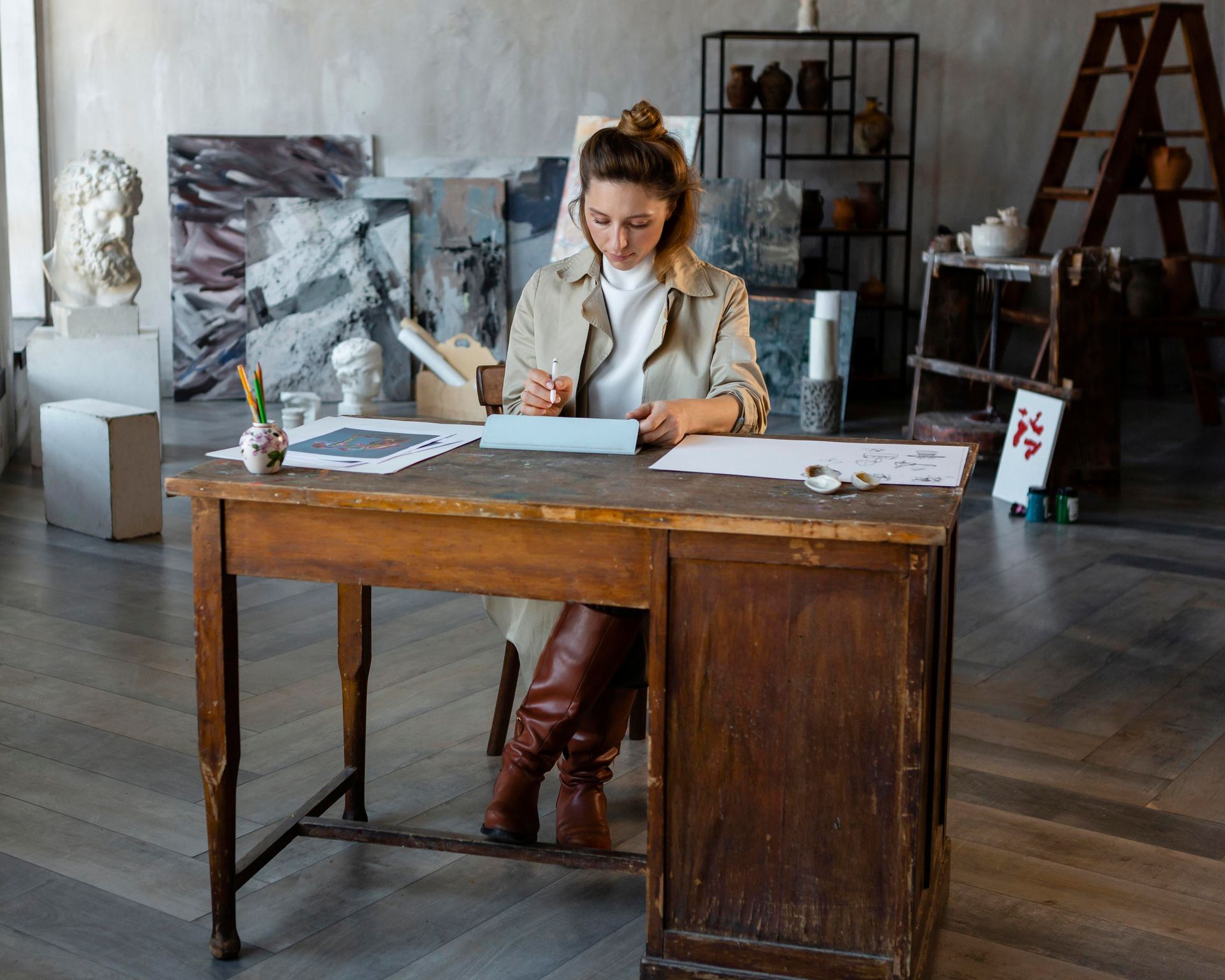 Woman in a tan coat and boots sits at a wooden desk in an art studio, drawing.